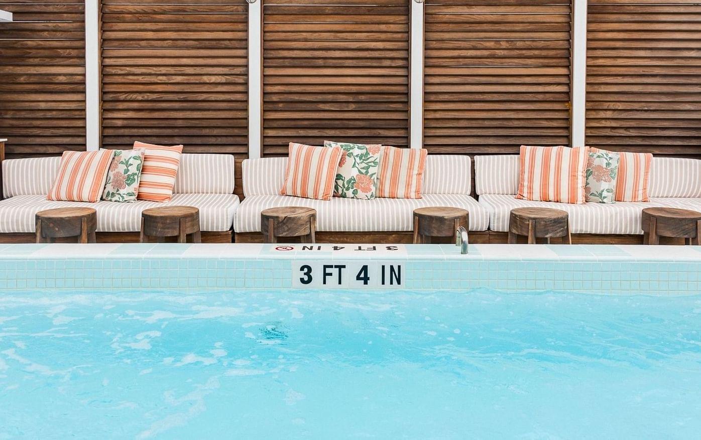 Striped and floral cushioned loungers along a wooden slatted wall beside a rooftop pool, with a depth marker reading 3 ft 4 in on the tile edge.