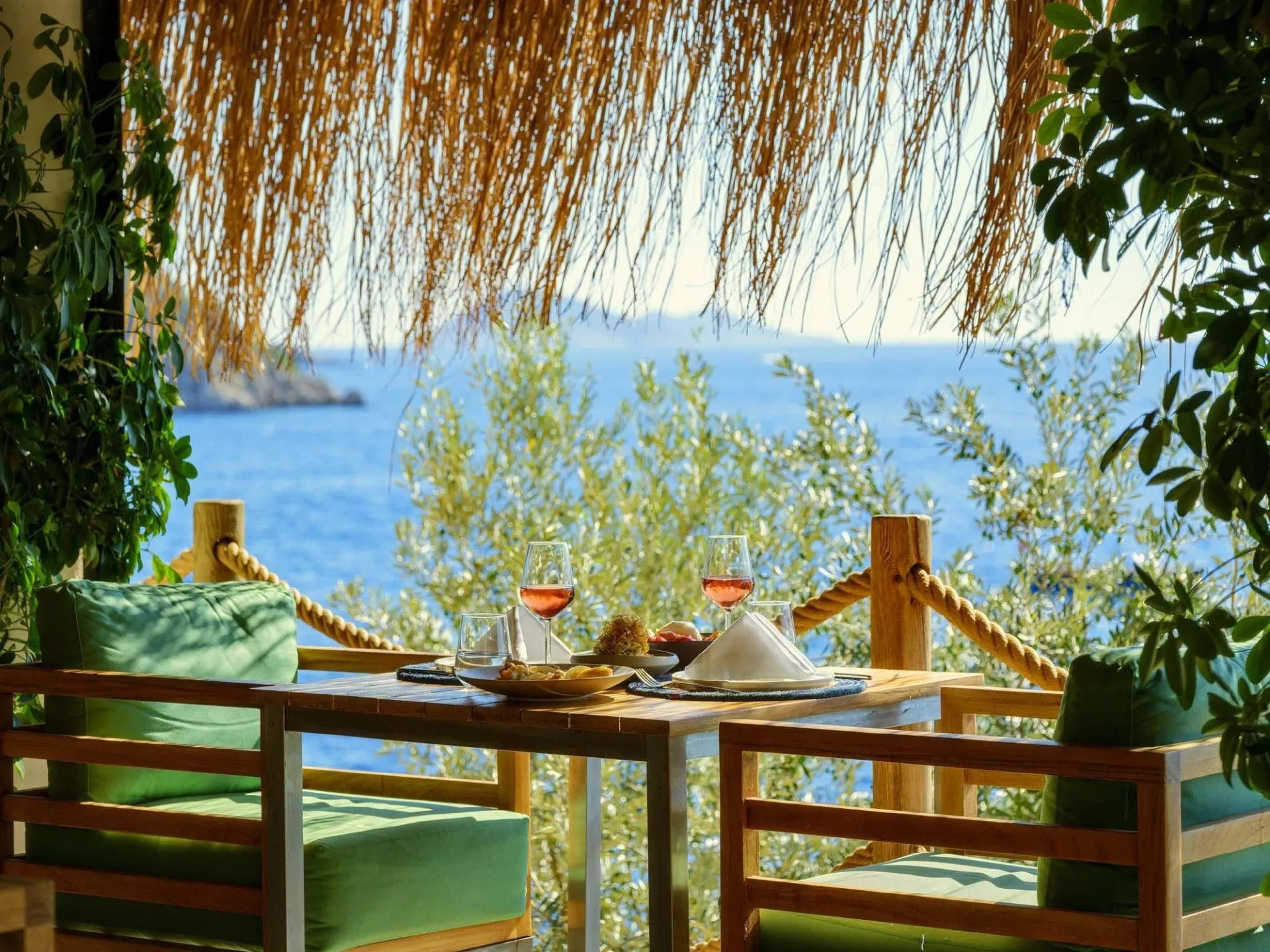 A table on a thatched terrace set with two glasses of rosé wine and small plates of food, with green-cushioned chairs and the sea and an olive tree behind.