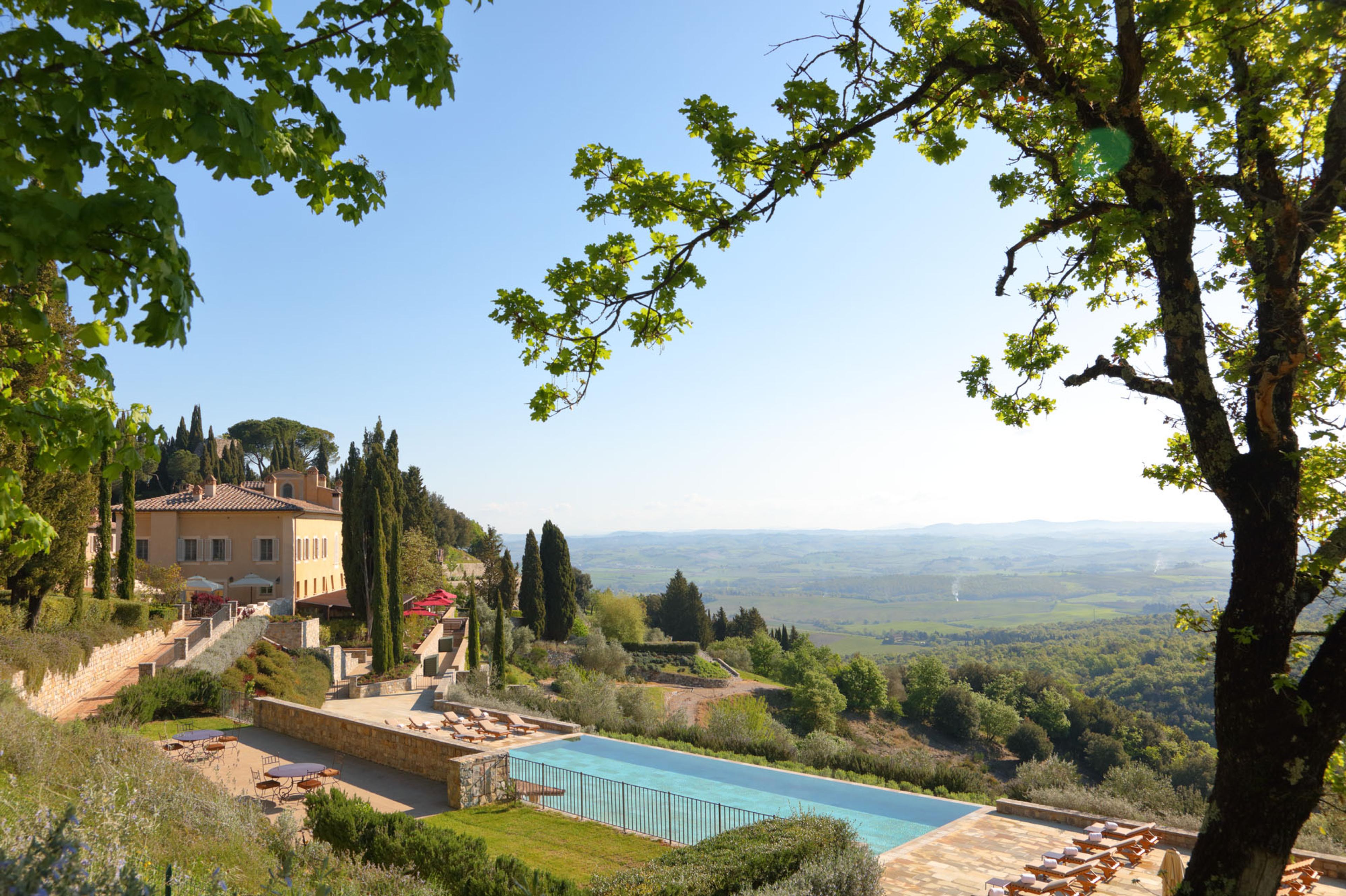 An infinity pool along a stone terrace below a cream stone villa, framed by overhanging tree branches, with a wide Tuscan valley beyond.
