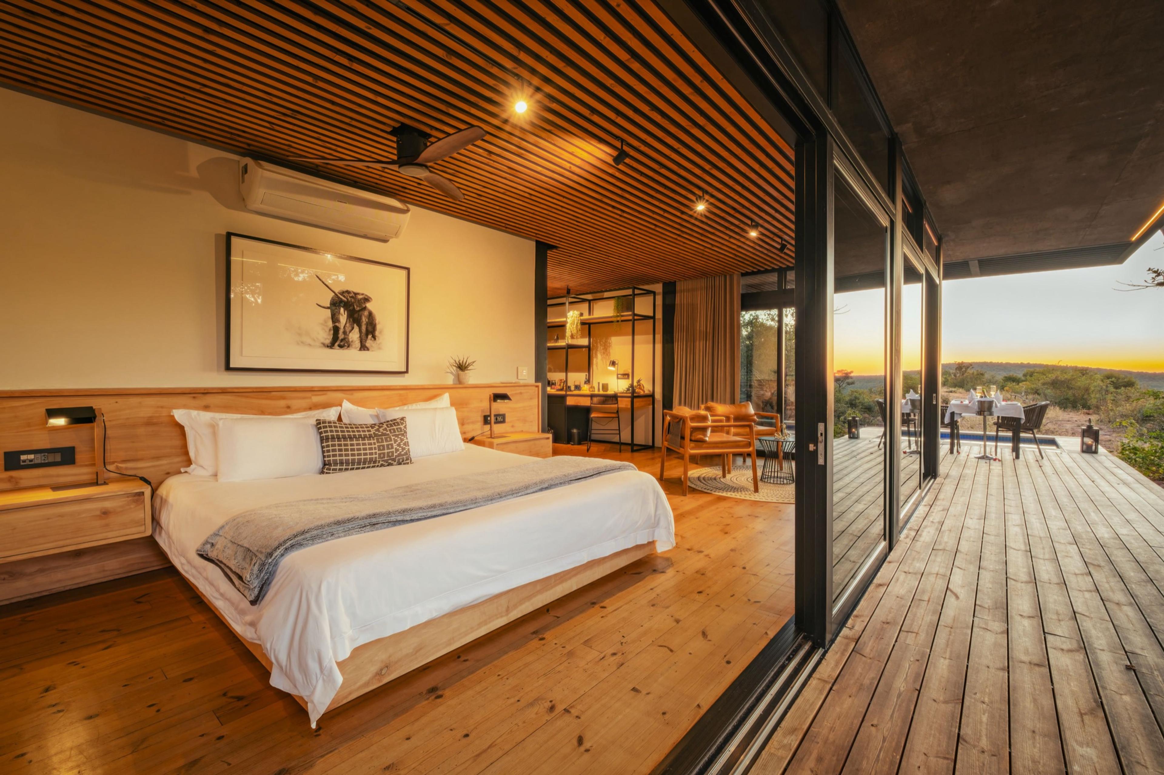 A bedroom with a wood-panelled ceiling and a made bed, its glass doors open onto a deck where a dining table is set for sunset.