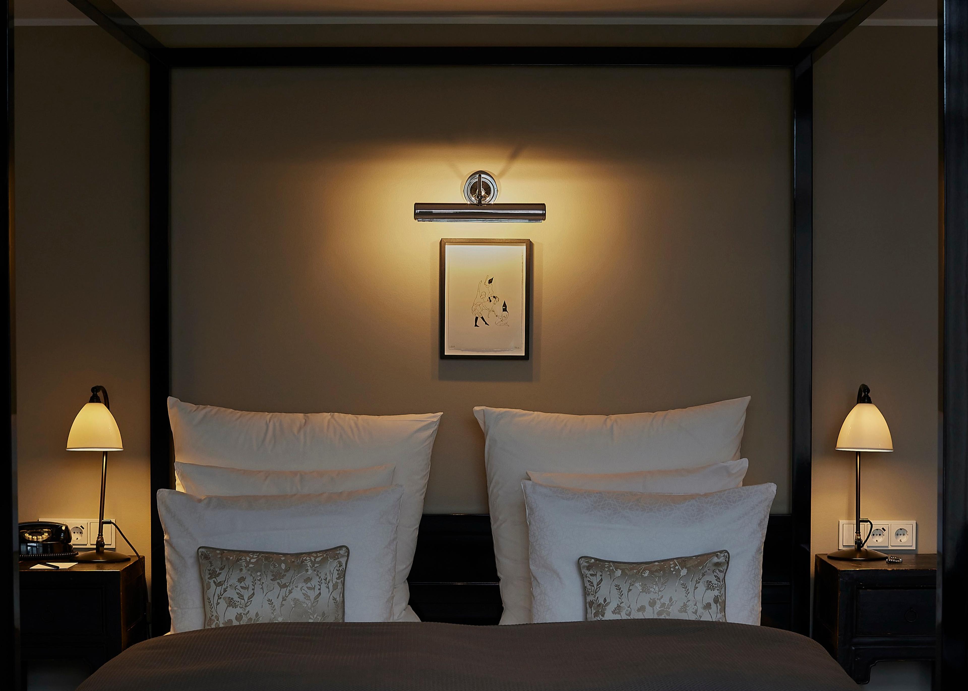 A bed with stacked white pillows and patterned cushions sits between two matching bedside tables, each with a black telephone and a lit lamp, under a small framed print.