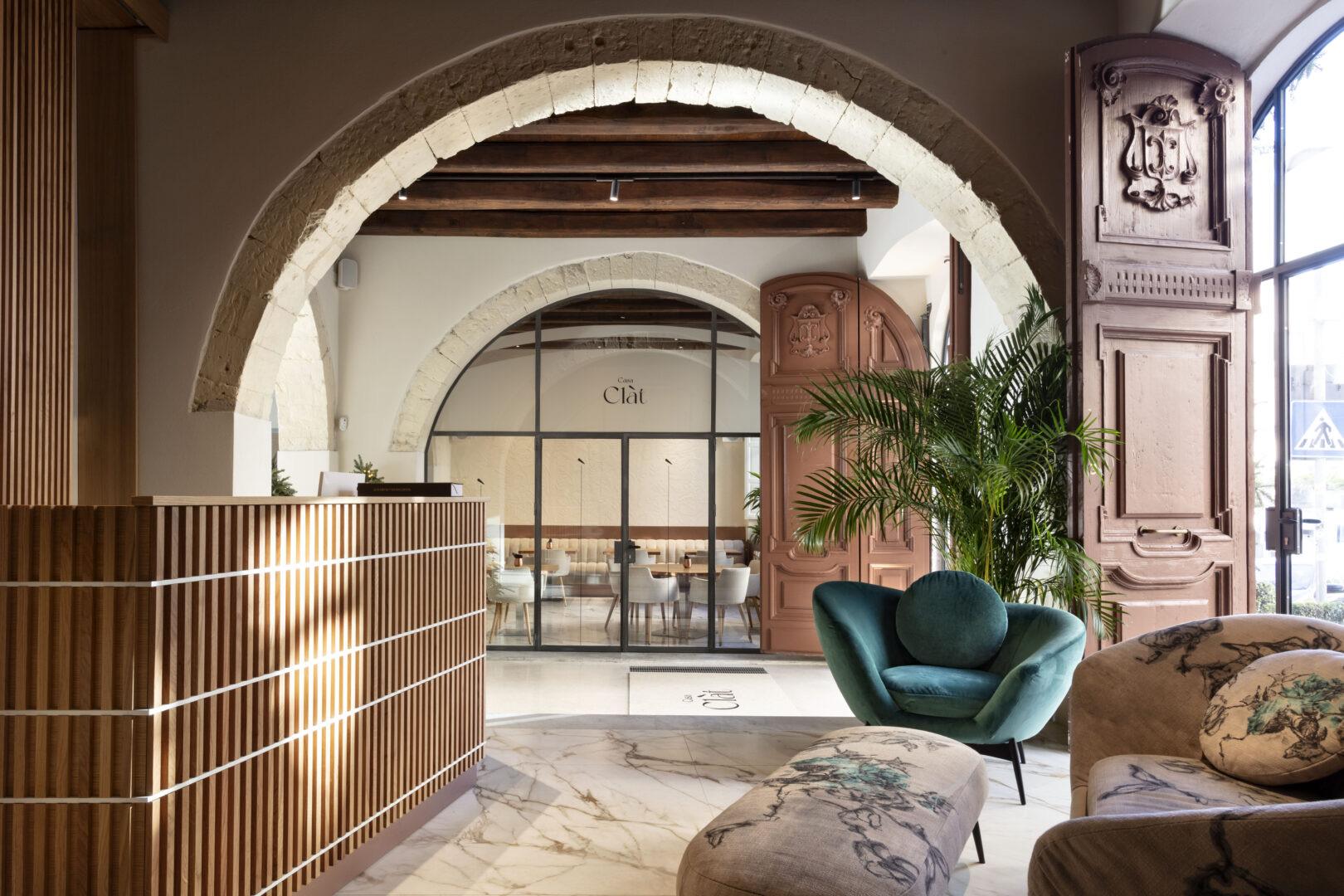 A hotel lobby with stone archways, a wood-slatted reception desk, a green velvet armchair, potted palms, and carved wooden doors opening to the street.