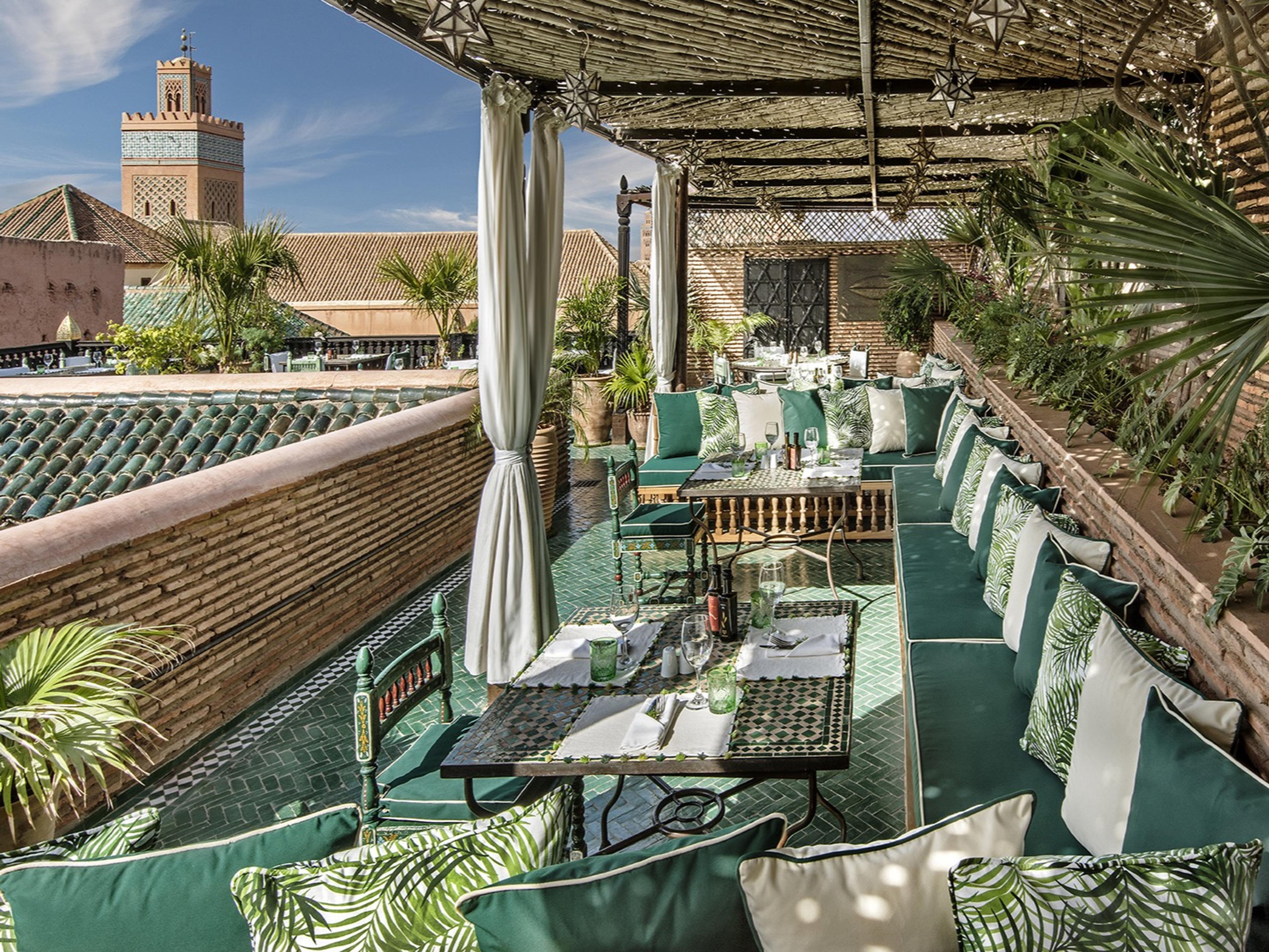 A rooftop terrace at La Sultana laid for lunch with green banquettes, looking over tiled rooftops to a minaret in the Kasbah