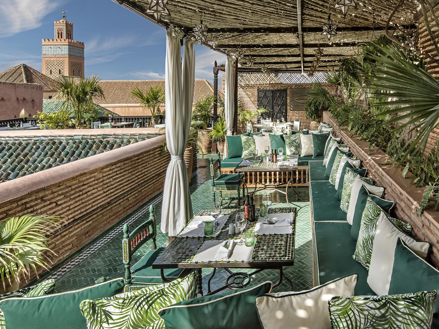 A rooftop terrace at La Sultana laid for lunch with green banquettes, looking over tiled rooftops to a minaret in the Kasbah
