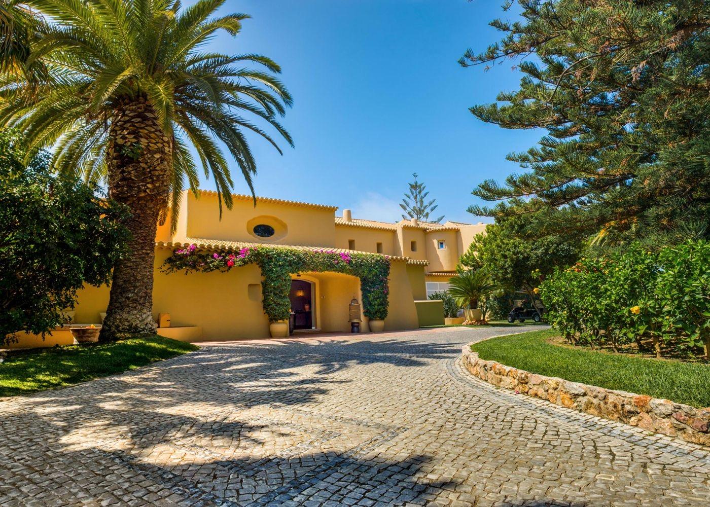 A cobbled driveway curves up to the ochre entrance of a hotel building framed by palm trees and bougainvillea, with a golf cart parked beyond.
