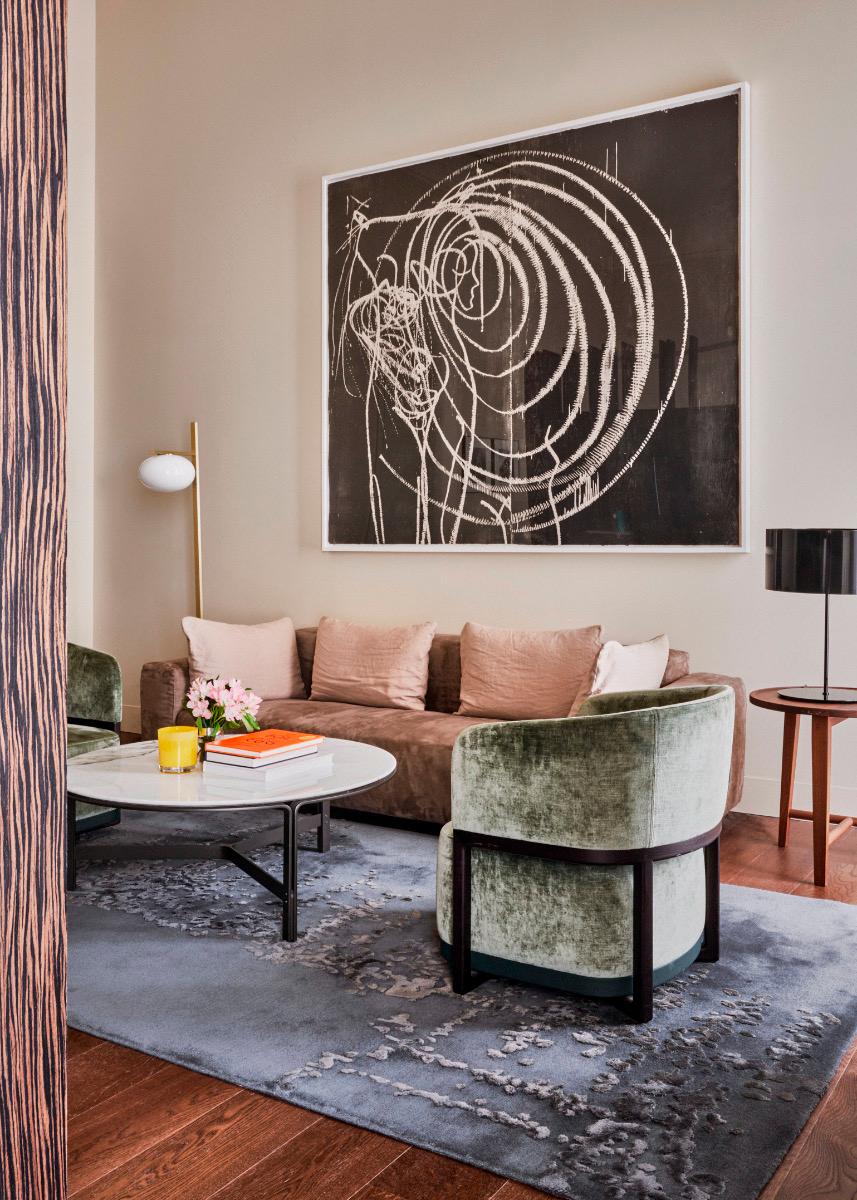 A brown velvet sofa and a green velvet armchair sit around a round marble coffee table with flowers and books, under a large abstract line drawing on the wall.