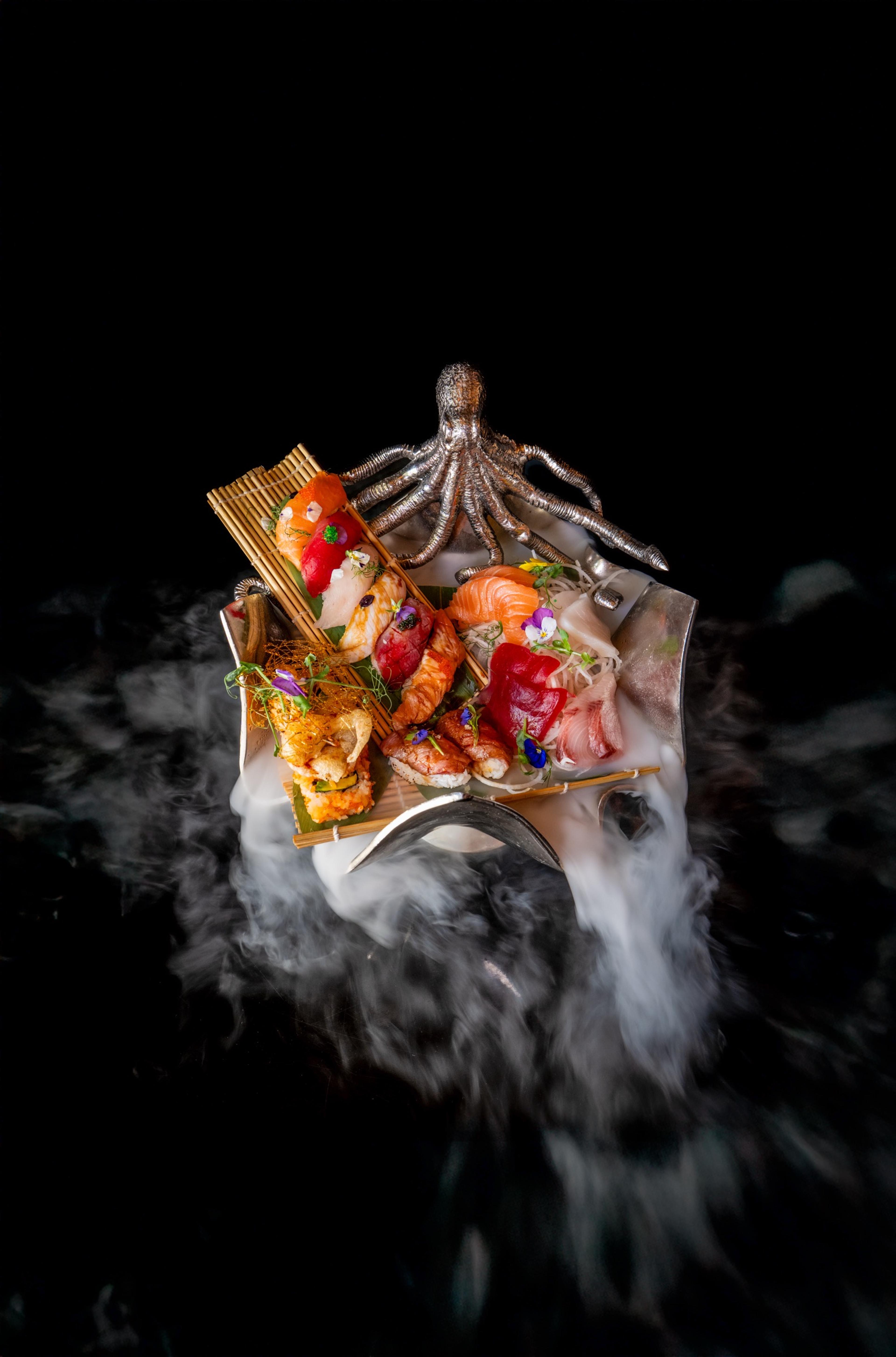 An overhead shot of thinly sliced grilled meat arranged on a plate with dots of sauce, steam or dry ice swirling around a silver octopus-shaped stand.