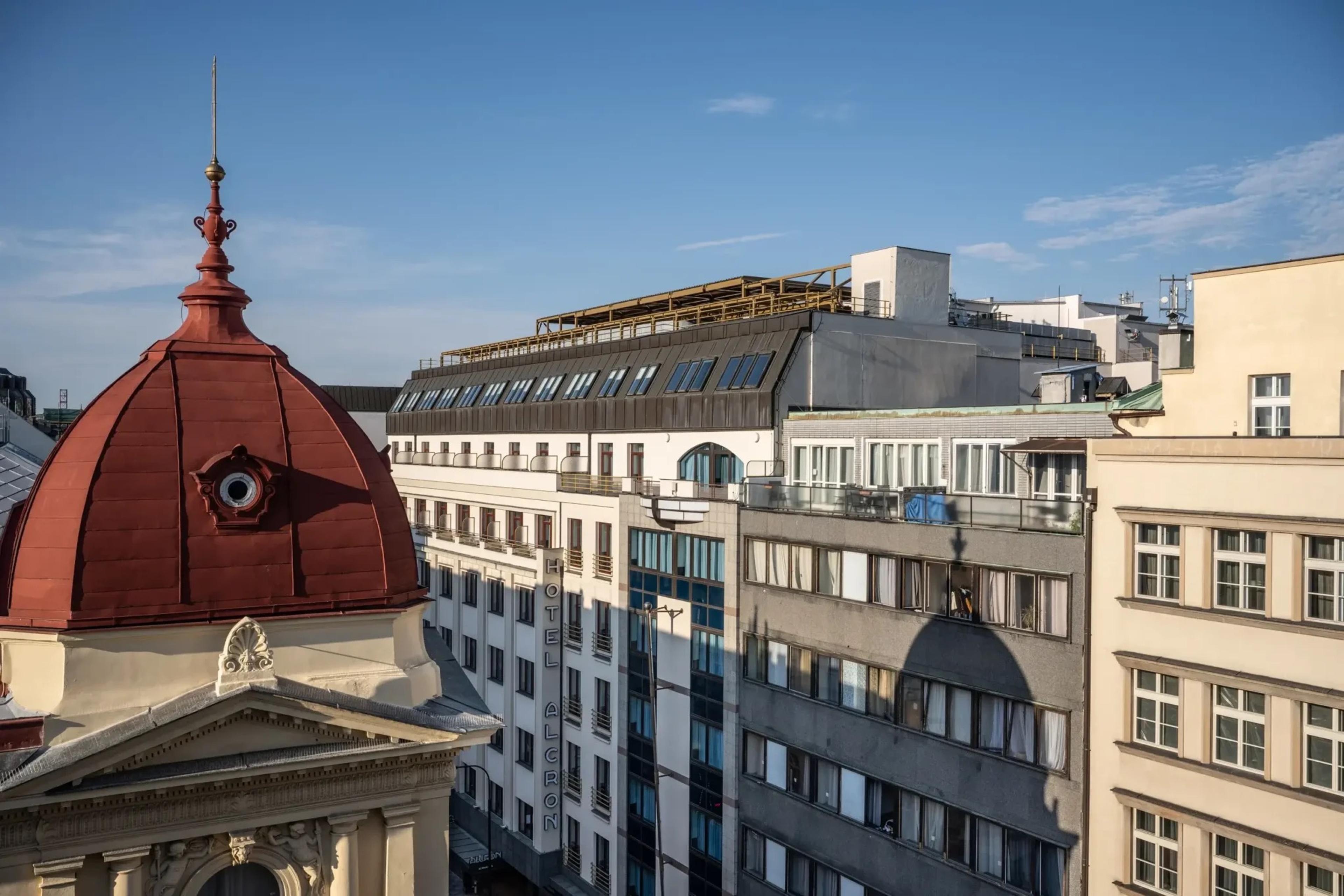 The hotel's facade seen from across a street, with a red copper dome in the foreground and a vertical HOTEL ALCRON sign on the building under a blue sky.