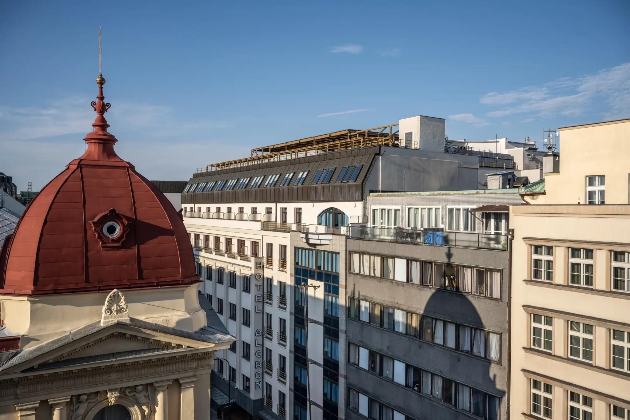 The hotel's facade seen from across a street, with a red copper dome in the foreground and a vertical HOTEL ALCRON sign on the building under a blue sky.