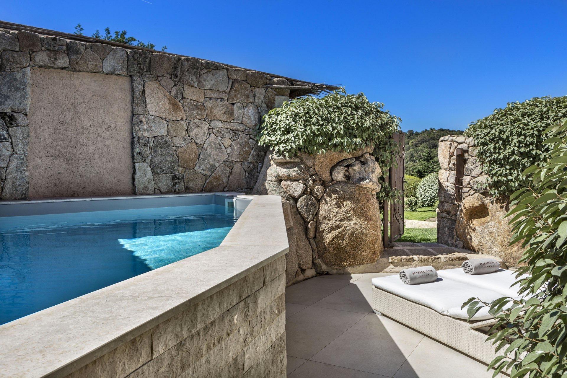 A small plunge pool set against a rough stone wall, with a daybed of rolled towels beside an archway leading to a garden.
