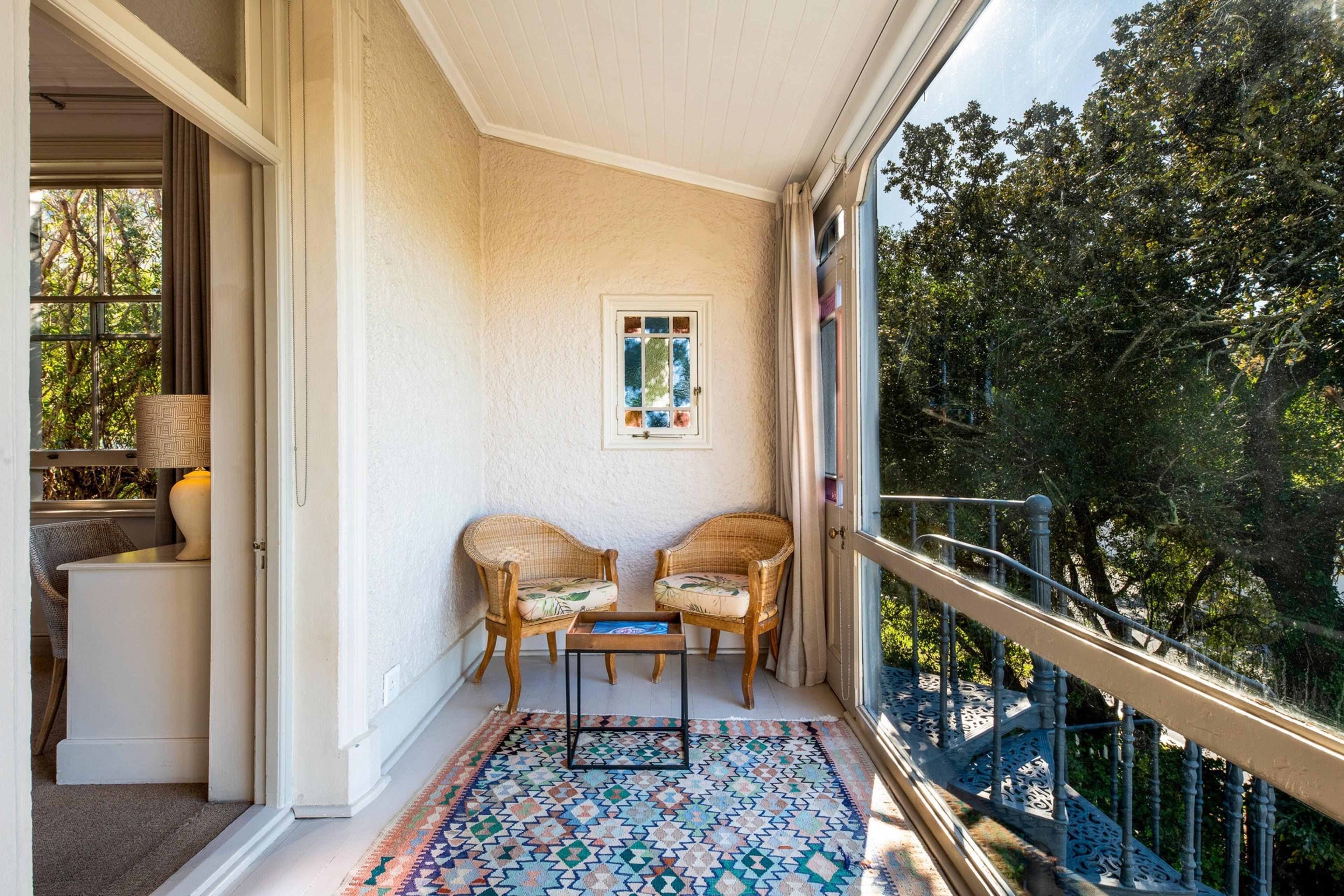 a narrow balcony with two wicker chairs around a low table on a patterned rug, screened by trees with an iron spiral staircase visible through the railing