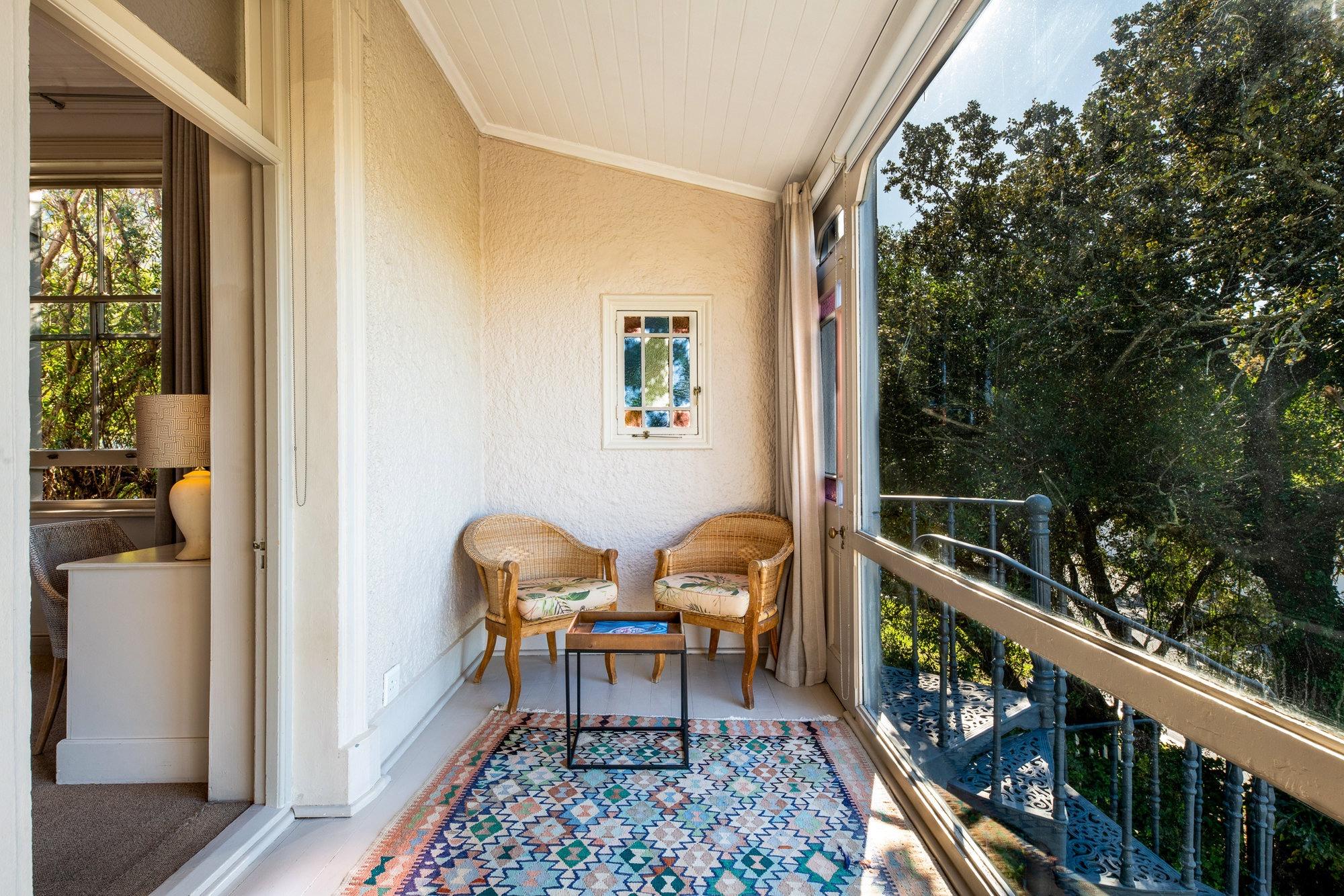 a narrow balcony with two wicker chairs around a low table on a patterned rug, screened by trees with an iron spiral staircase visible through the railing