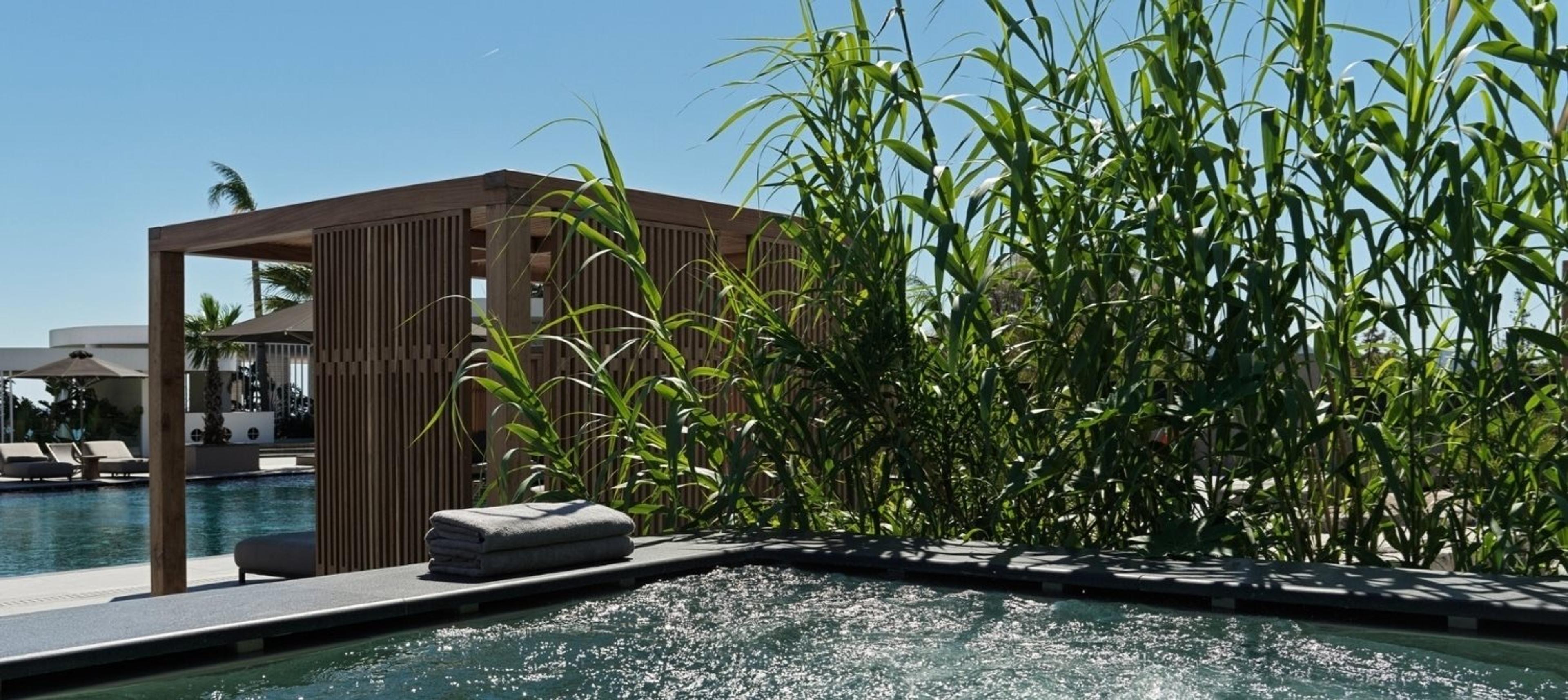 Rolled towels beside a wooden spa pavilion, tall grasses in the foreground and a plunge pool in the corner.