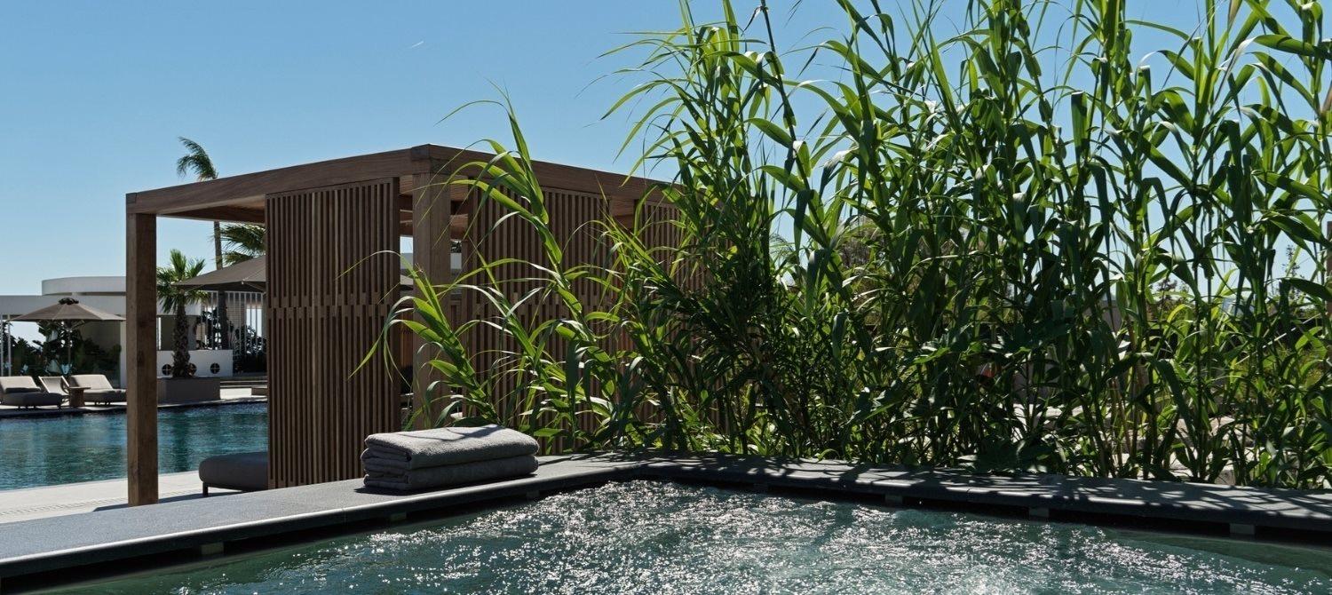 Rolled towels beside a wooden spa pavilion, tall grasses in the foreground and a plunge pool in the corner.