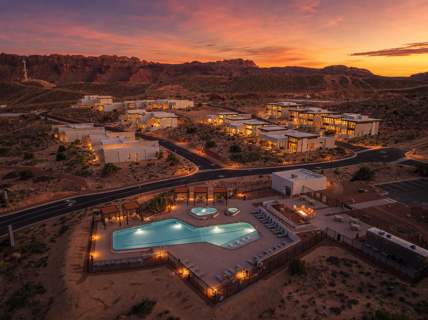 An aerial view at sunset of a desert resort with casitas, a lit pool and hot tubs, roads, and red rock cliffs beyond.
