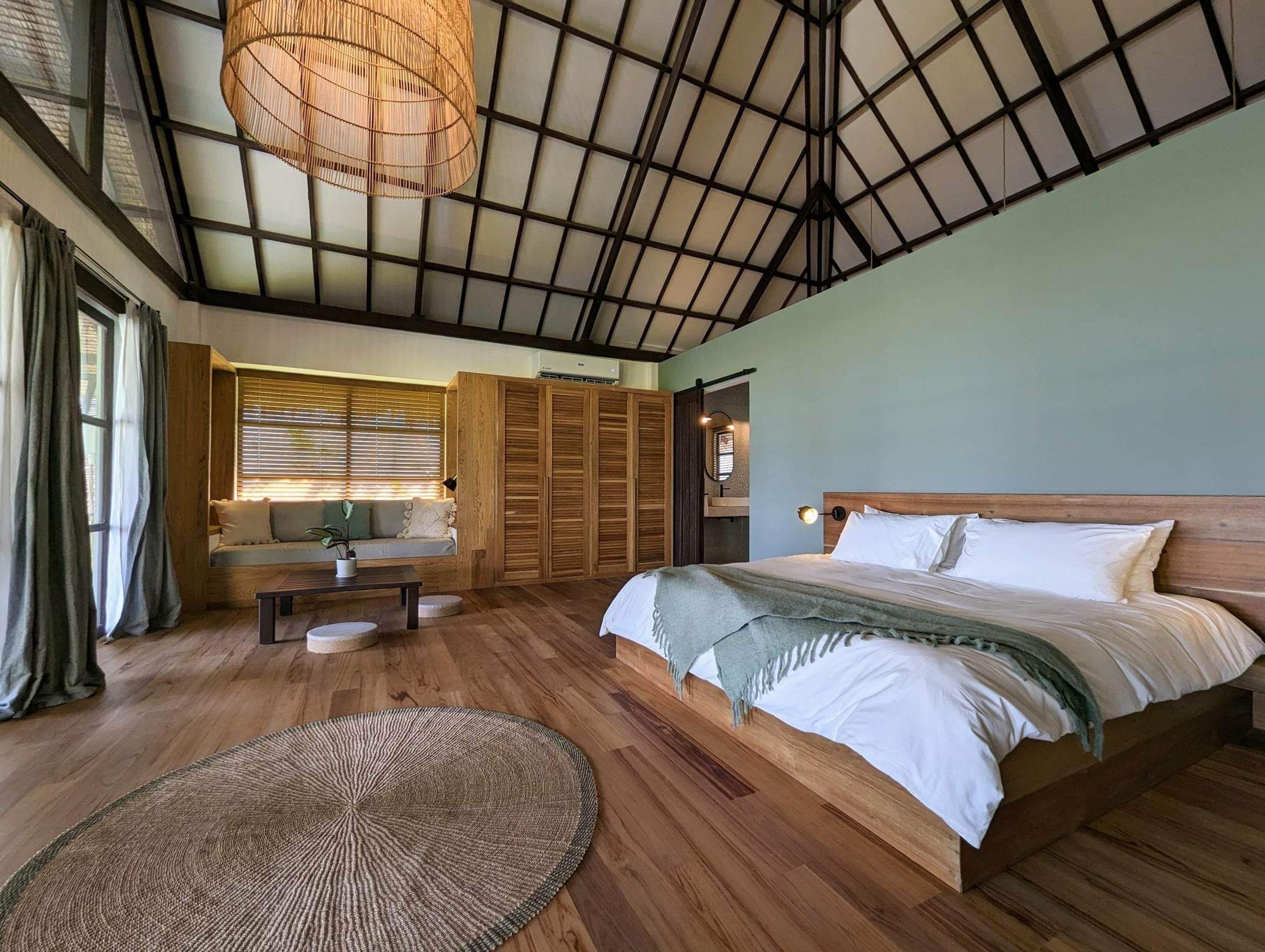 A bedroom with a wood-framed bed against a sage green wall, under a high vaulted ceiling with a woven pendant light.