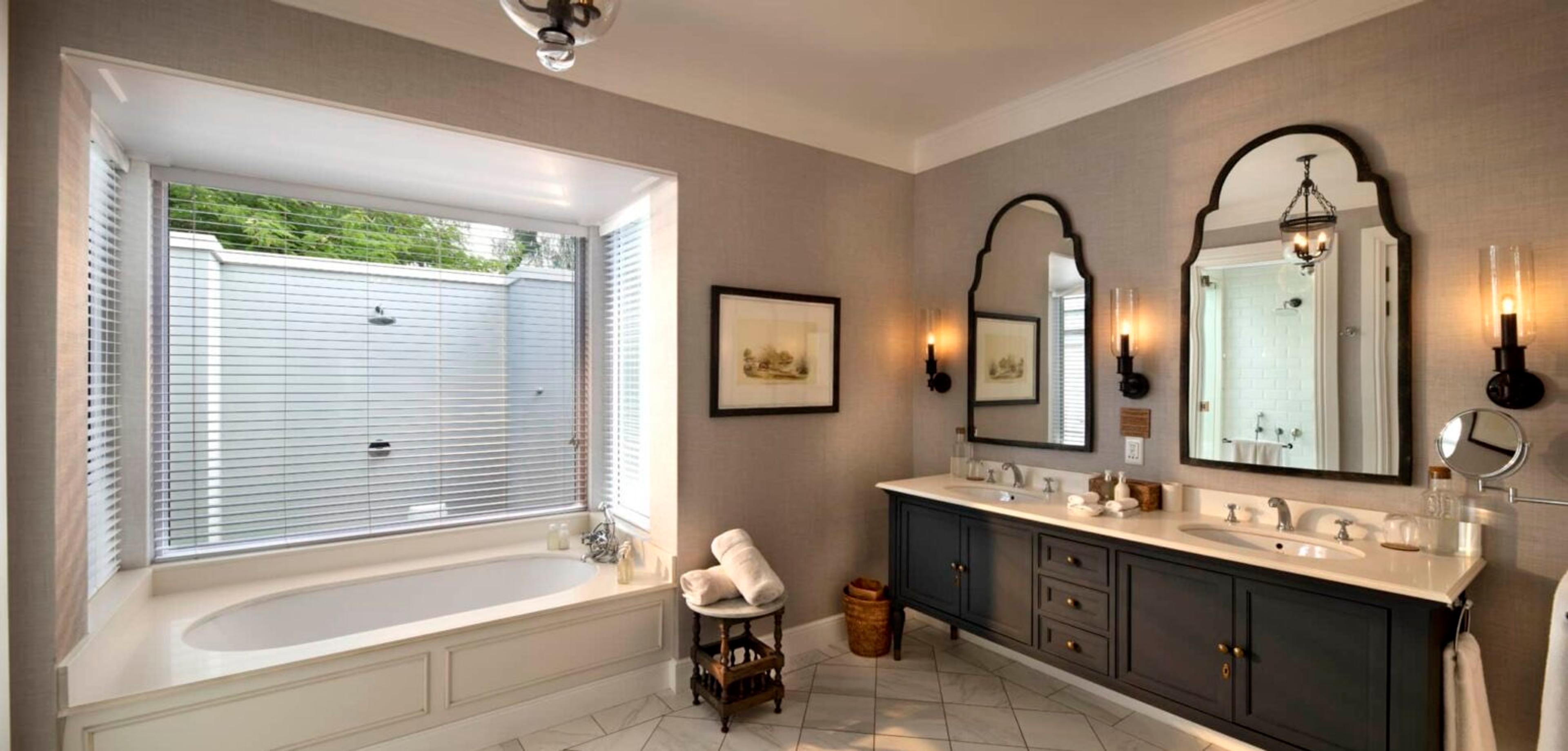 A freestanding bath under a bay window beside a double vanity with two arched mirrors.