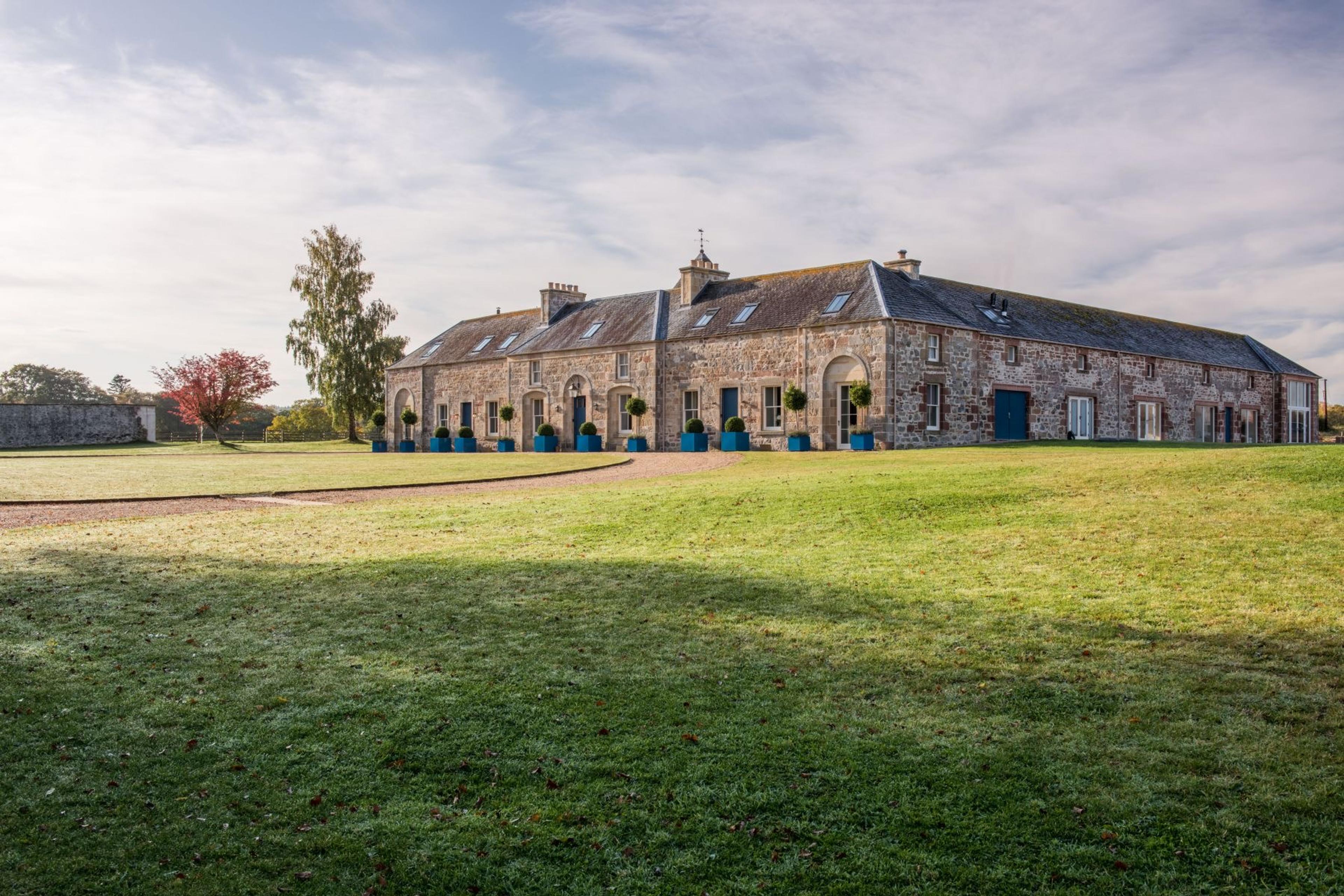 A long stone steading with slate roofs and blue potted topiaries along its front, seen across an open lawn under a pale sky.