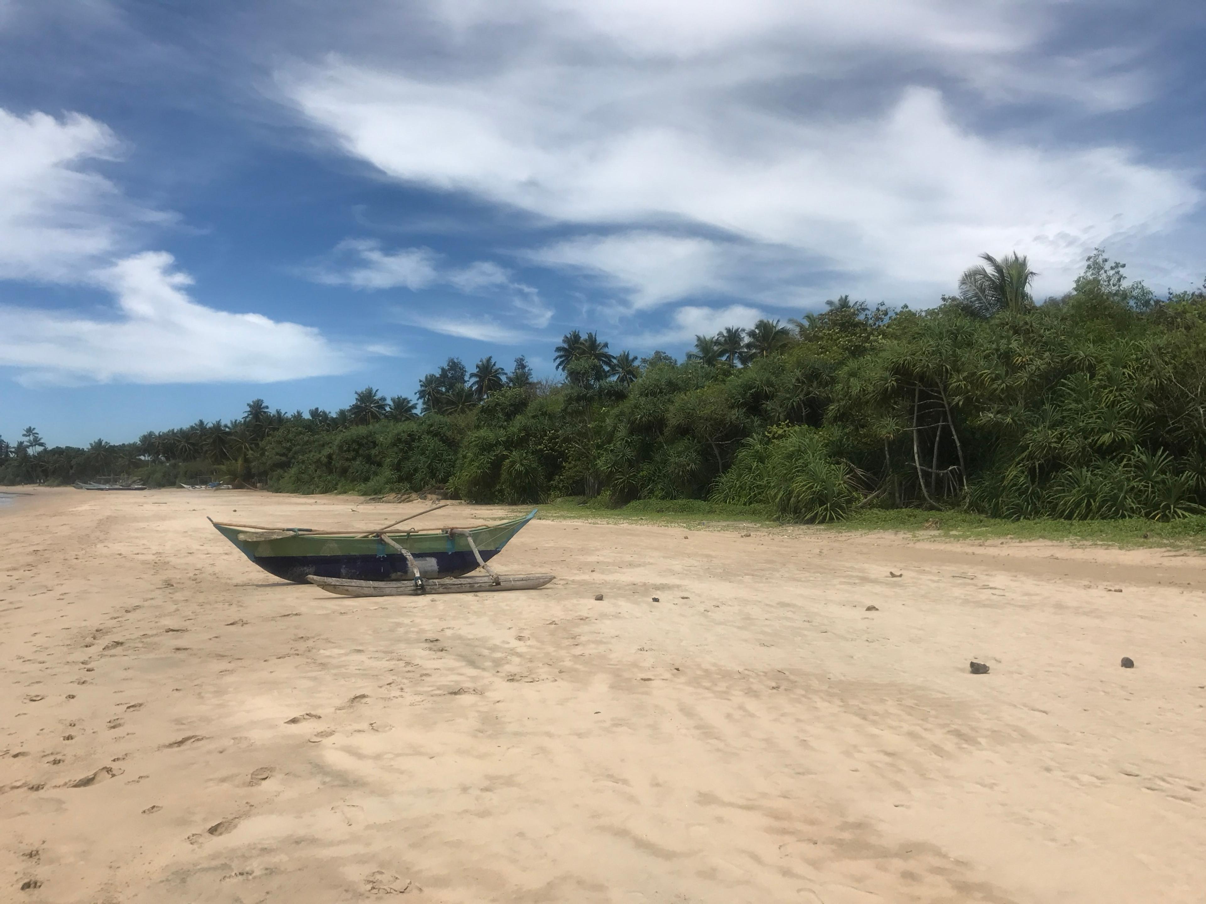 A wooden outrigger fishing boat drawn up on an empty sand beach at Mawella, with dense palms and jungle behind under a blue sky