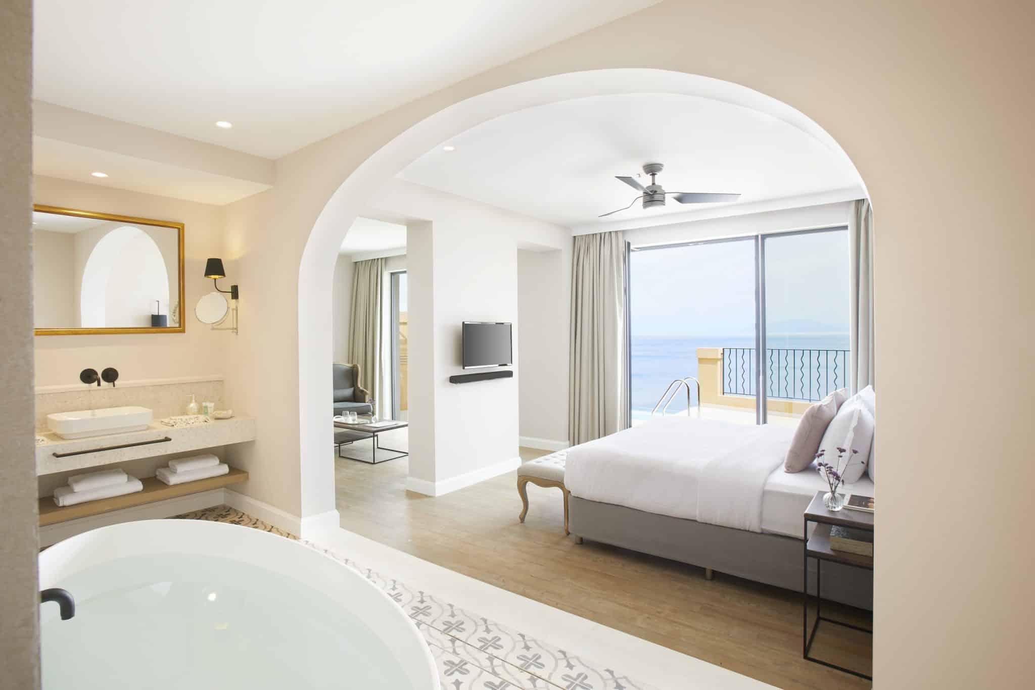 A free-standing bathtub and vanity in the foreground, seen through a stone archway into a bedroom with a bed facing a glass door and sea view.