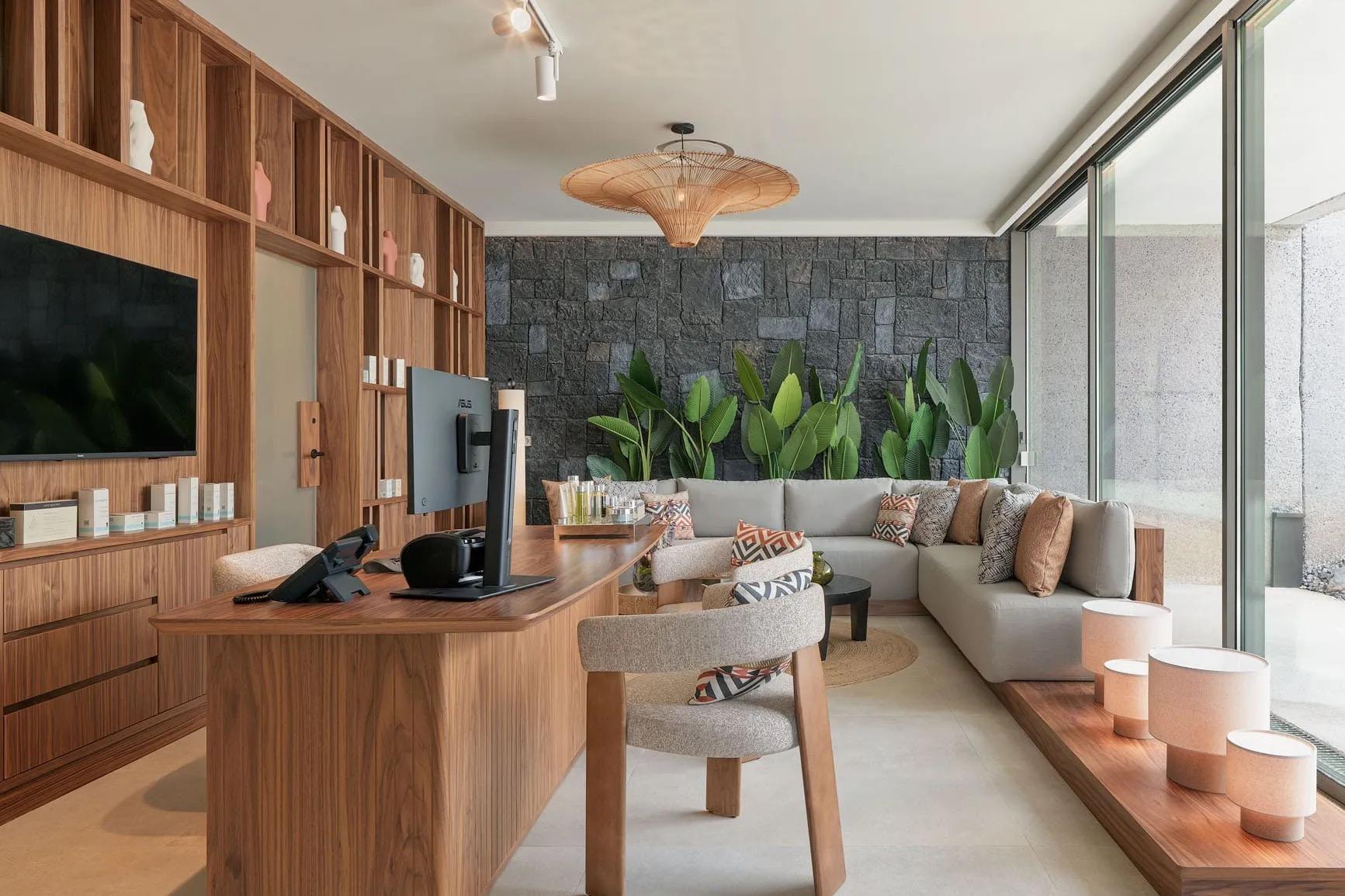 A lounge area with a grey sectional sofa, patterned cushions, a wall of dark stone, and a wooden desk with a monitor.