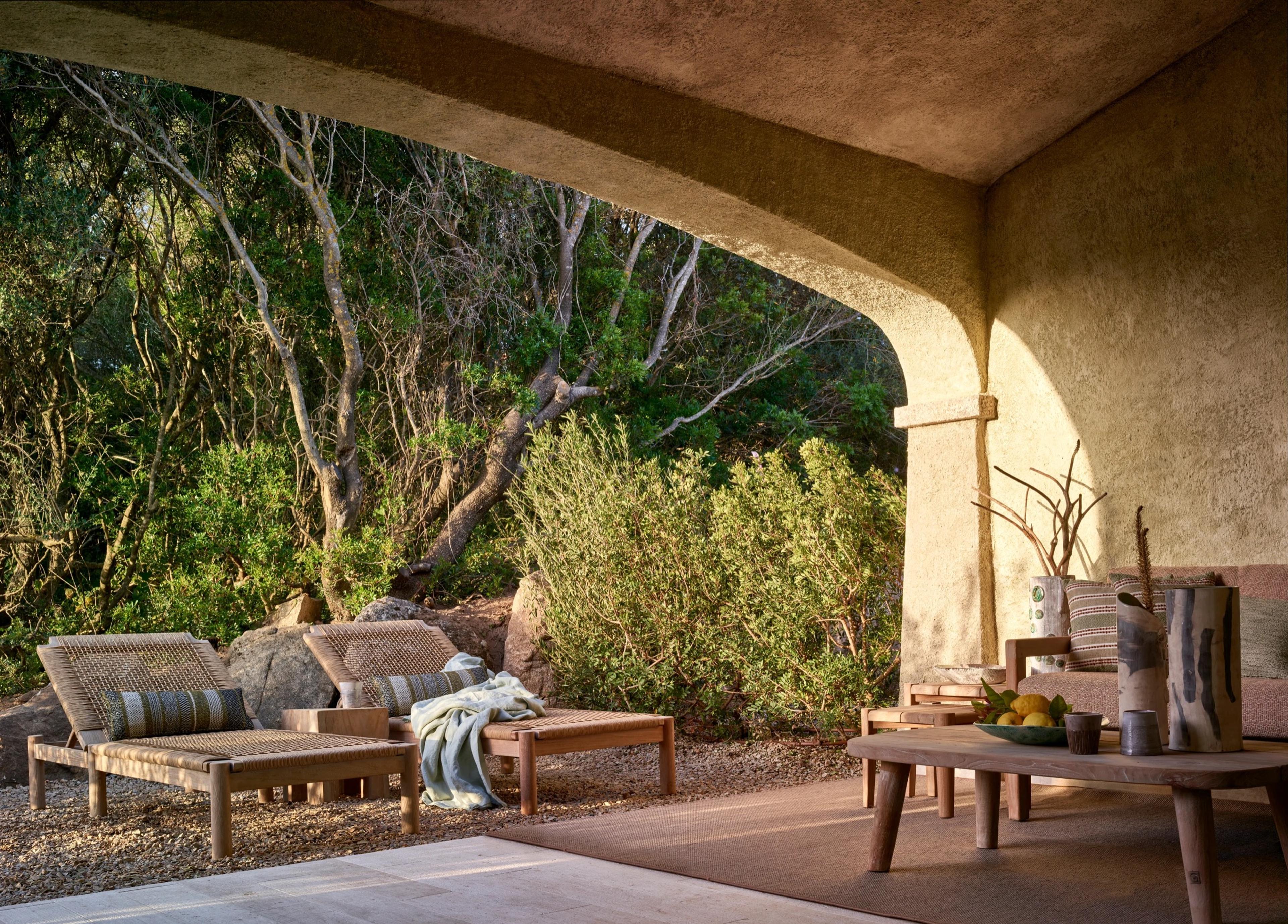 a covered stone terrace with two woven sun loungers facing trees and boulders, lit by low evening light