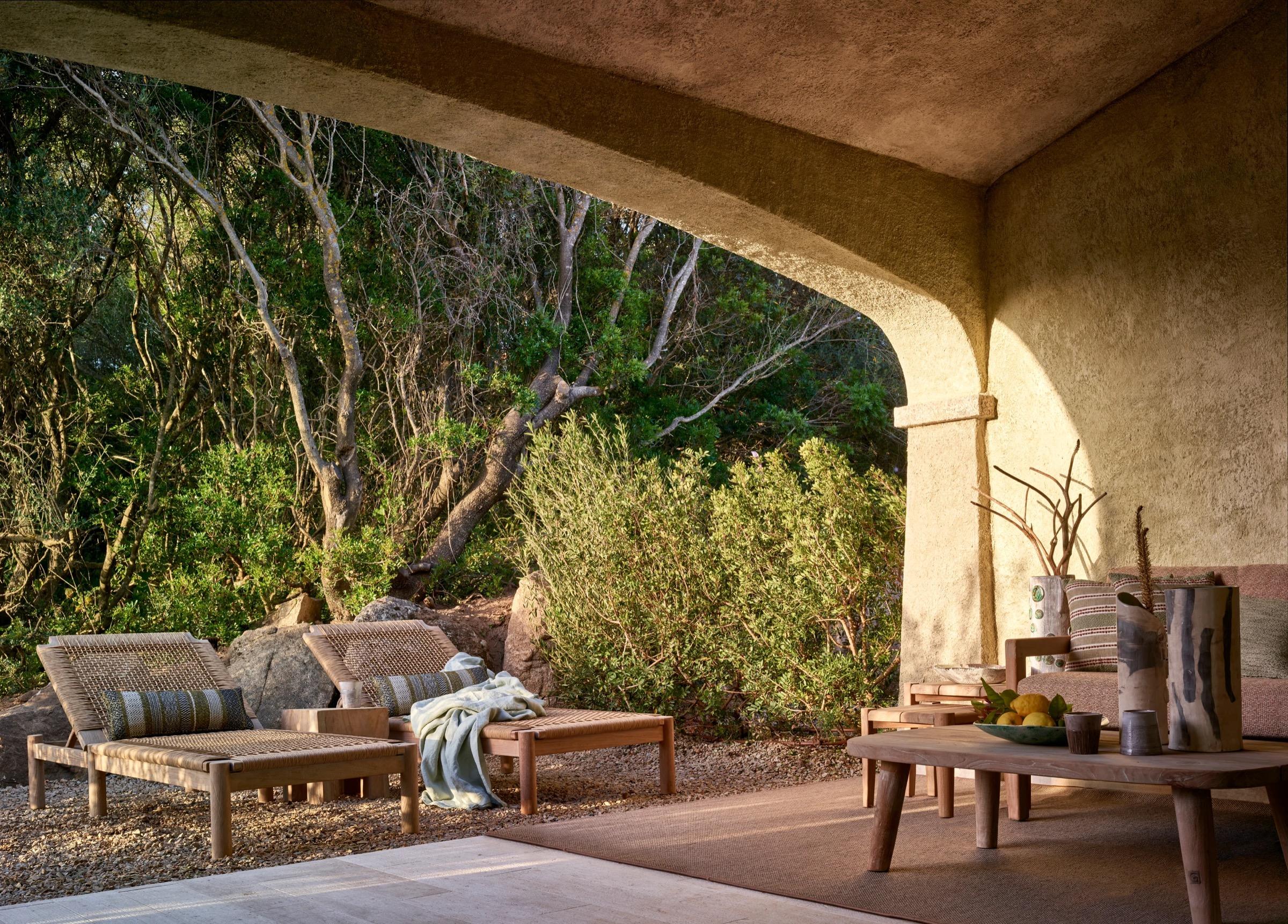 a covered stone terrace with two woven sun loungers facing trees and boulders, lit by low evening light
