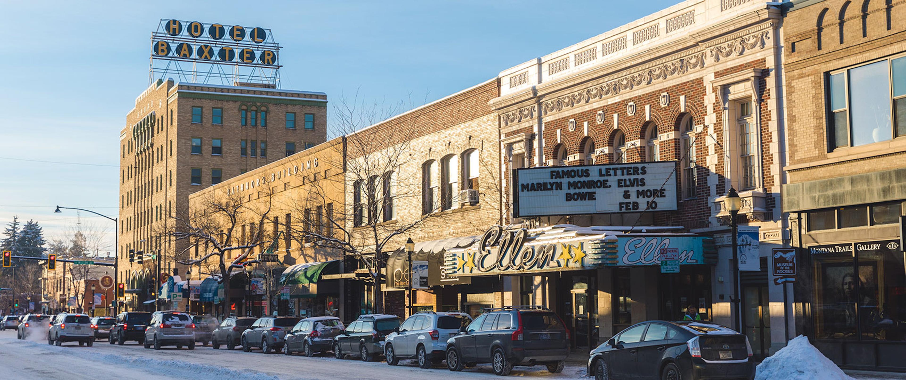 A snowy downtown street lined with parked cars and historic brick buildings, with the Hotel Baxter sign and a cinema marquee lit against the evening sky.