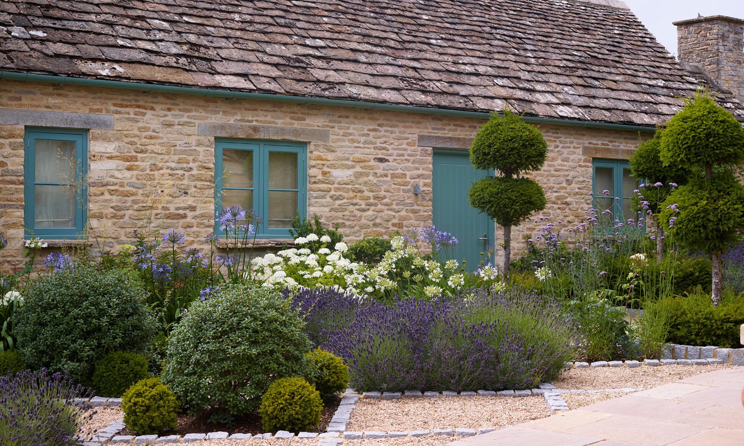 A honey-coloured stone cottage with teal sash windows and a matching door, fronted by a bed of agapanthus, hydrangeas and lavender.