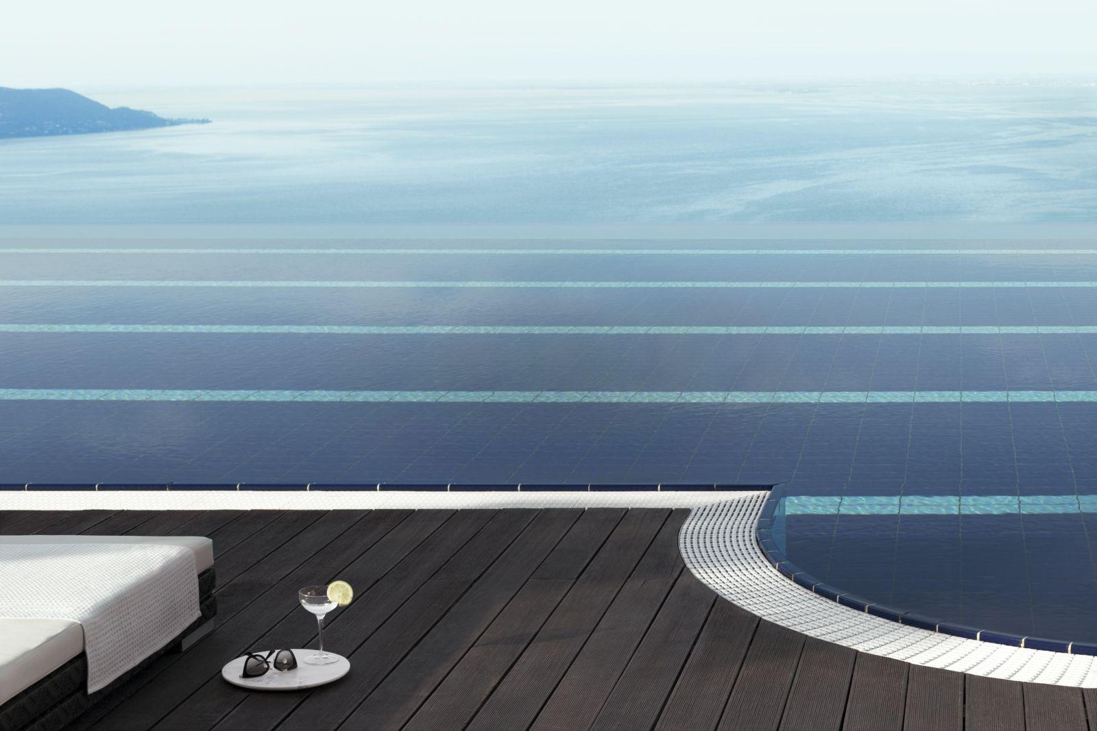 The tiled edge of an infinity pool with a lounger, a folded towel, and a cocktail glass and sunglasses on a round tray, overlooking the lake.