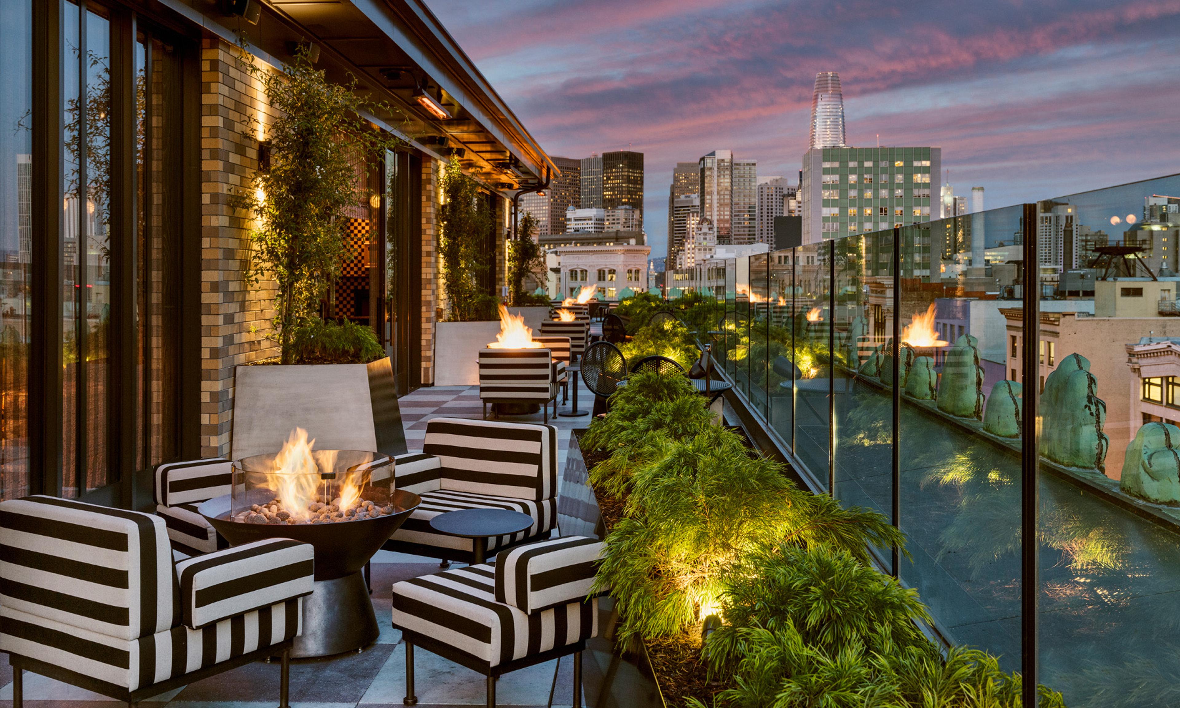 A rooftop terrace at dusk with striped armchairs around a lit fire pit, a glass railing, and the San Francisco skyline under a pink sky.