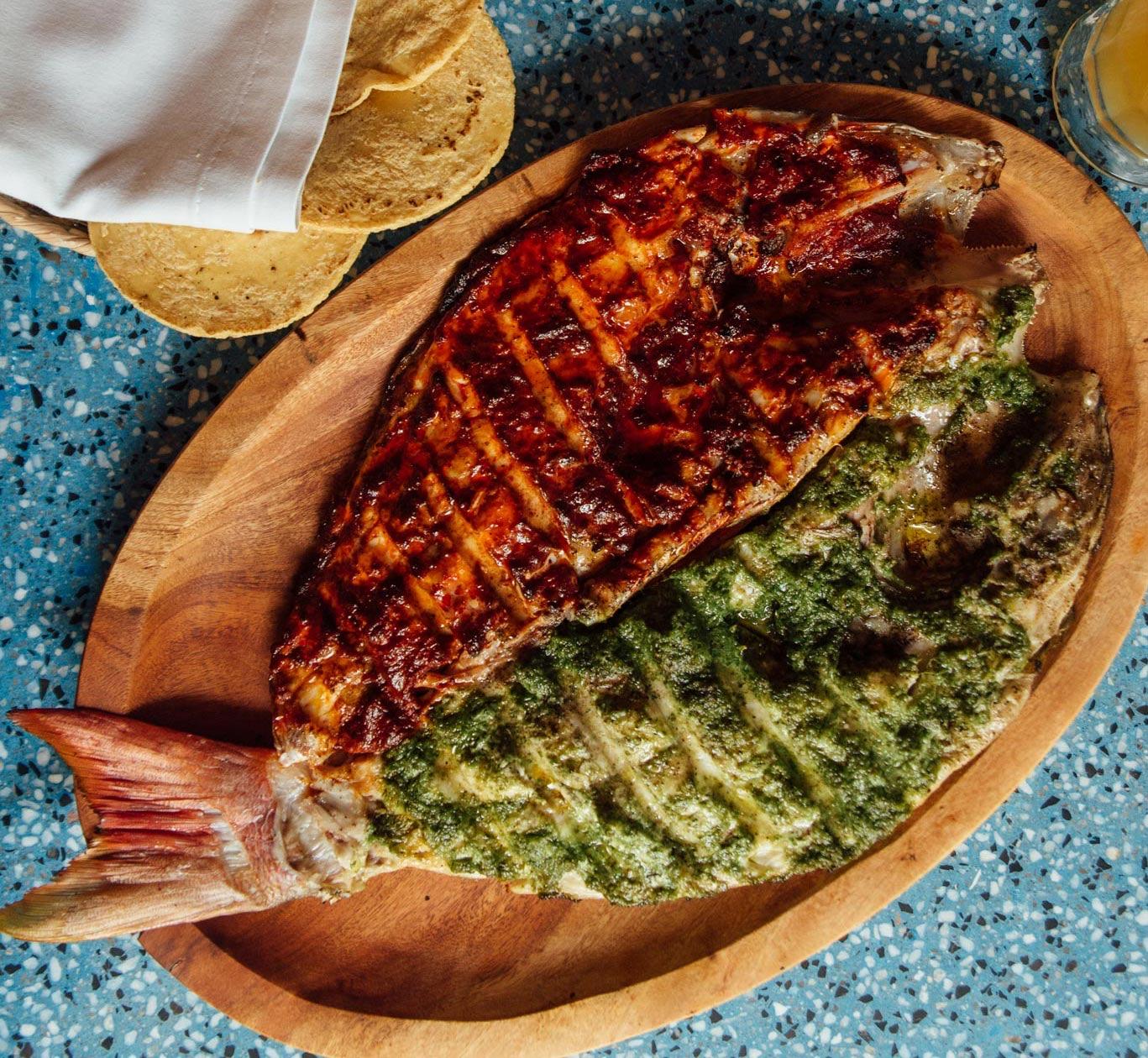 A whole grilled fish, butterflied and painted half red and half green, on a wooden board at Contramar