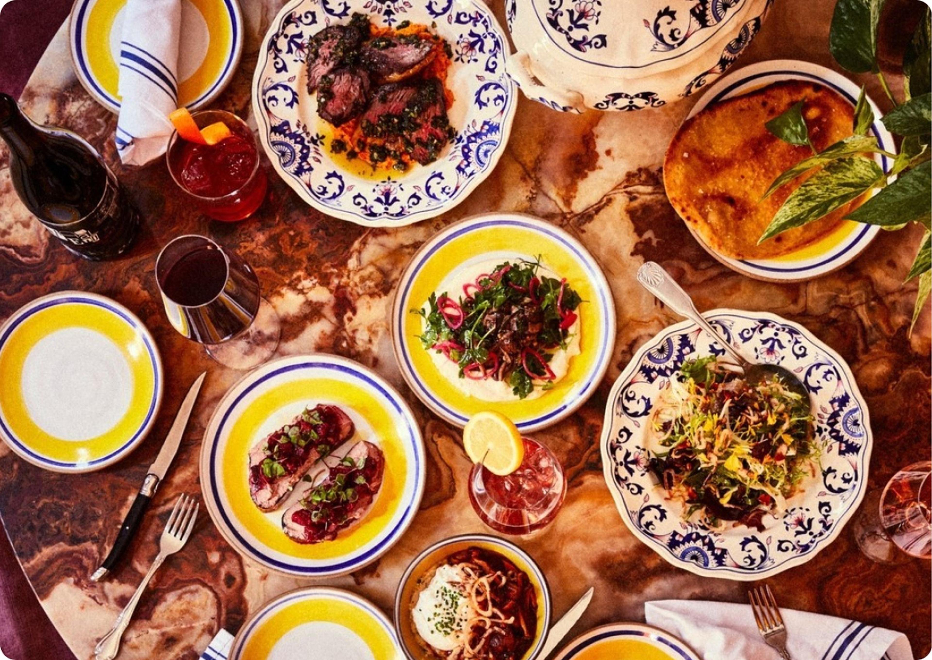 An overhead view of a marble table set with small plates of seared meat, flatbread and salads, alongside two cocktails, a glass of red wine and a glass of white.