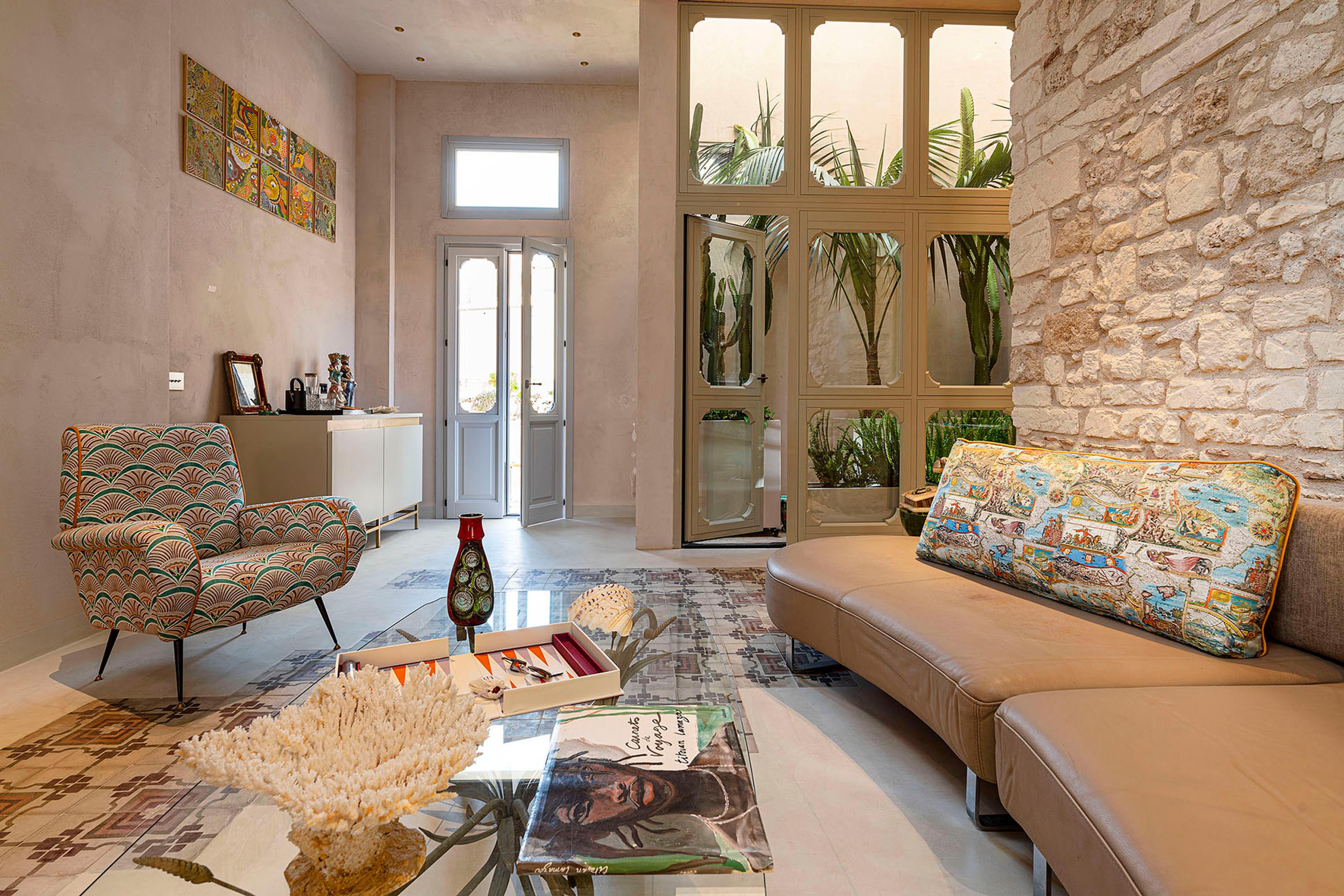 A living room with an exposed stone wall, a curved leather sofa, a patterned armchair, and glass doors opening onto a plant filled courtyard.