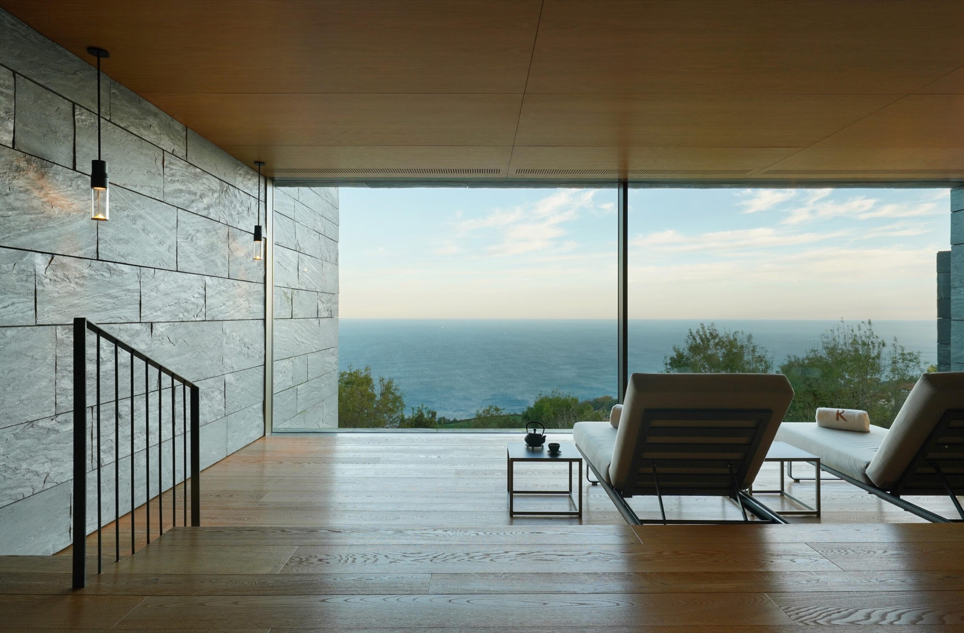 Two sunbeds beside a low table inside a stone-walled spa room, facing a full-height window onto the sea and trees.