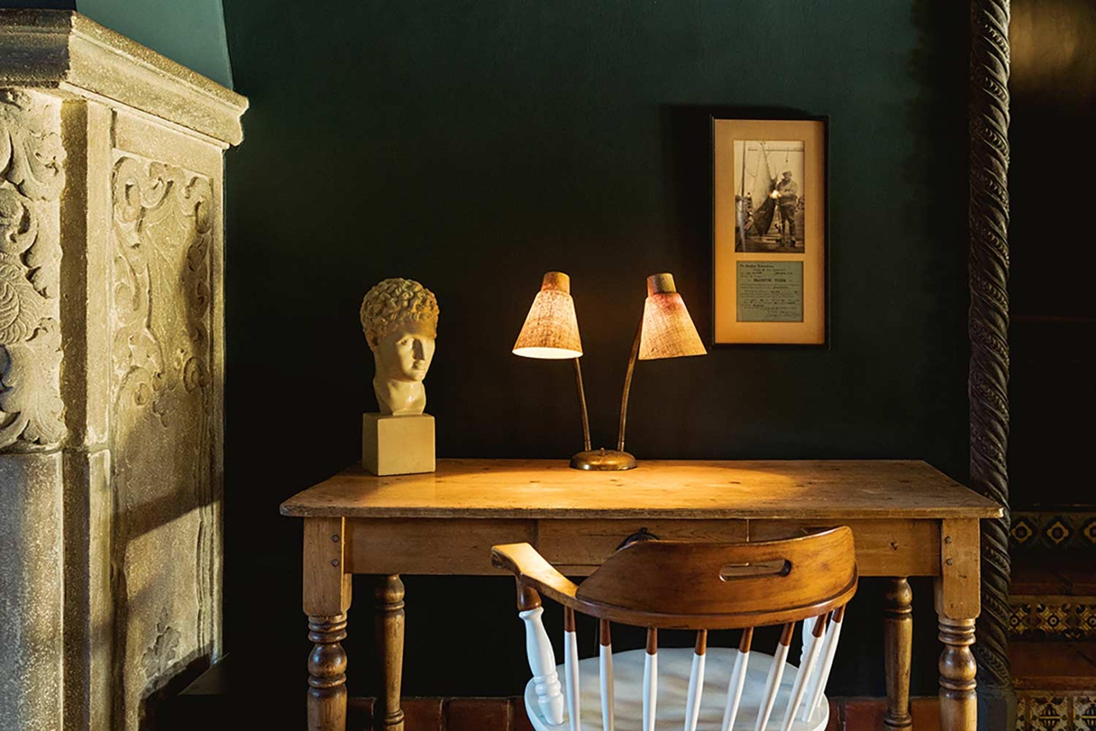 A plaster bust on a wooden desk beside two reading lamps and a small framed photograph, against a dark green wall and a carved stone chimneypiece.