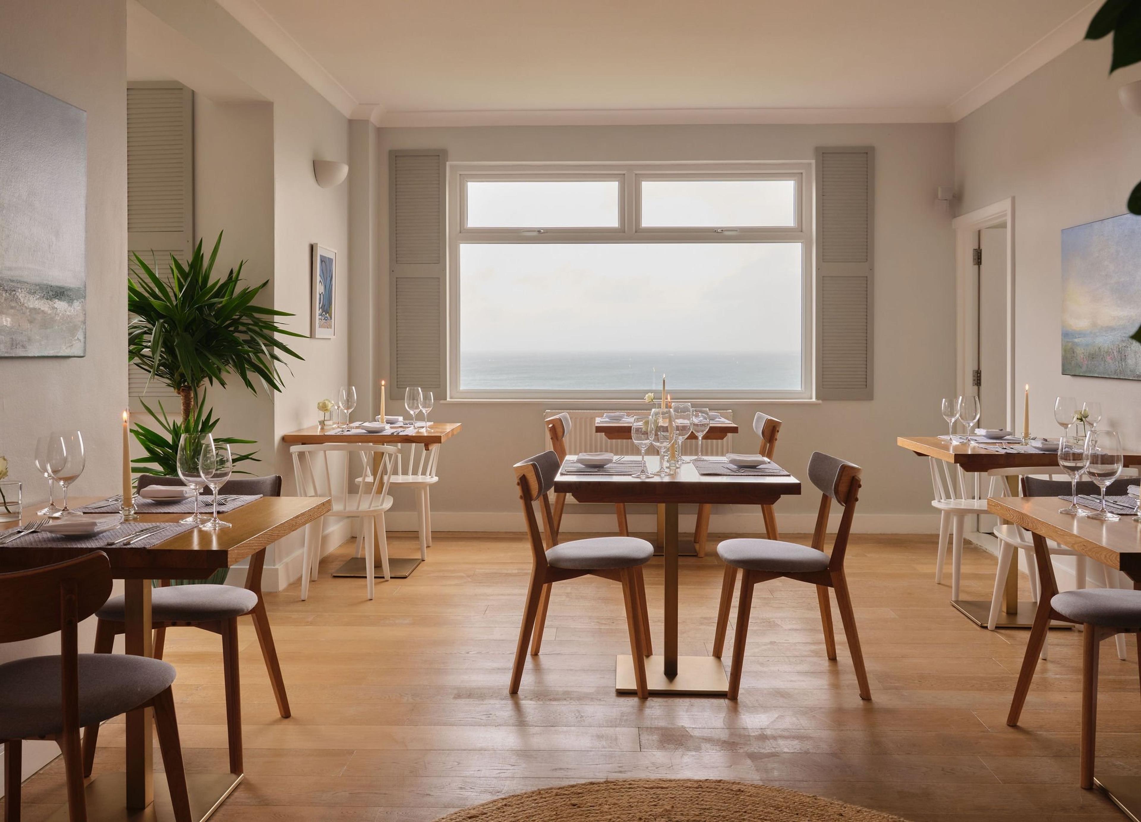 A restaurant dining room with wooden tables set for dinner, candles lit, and a wide window looking straight out to the sea.