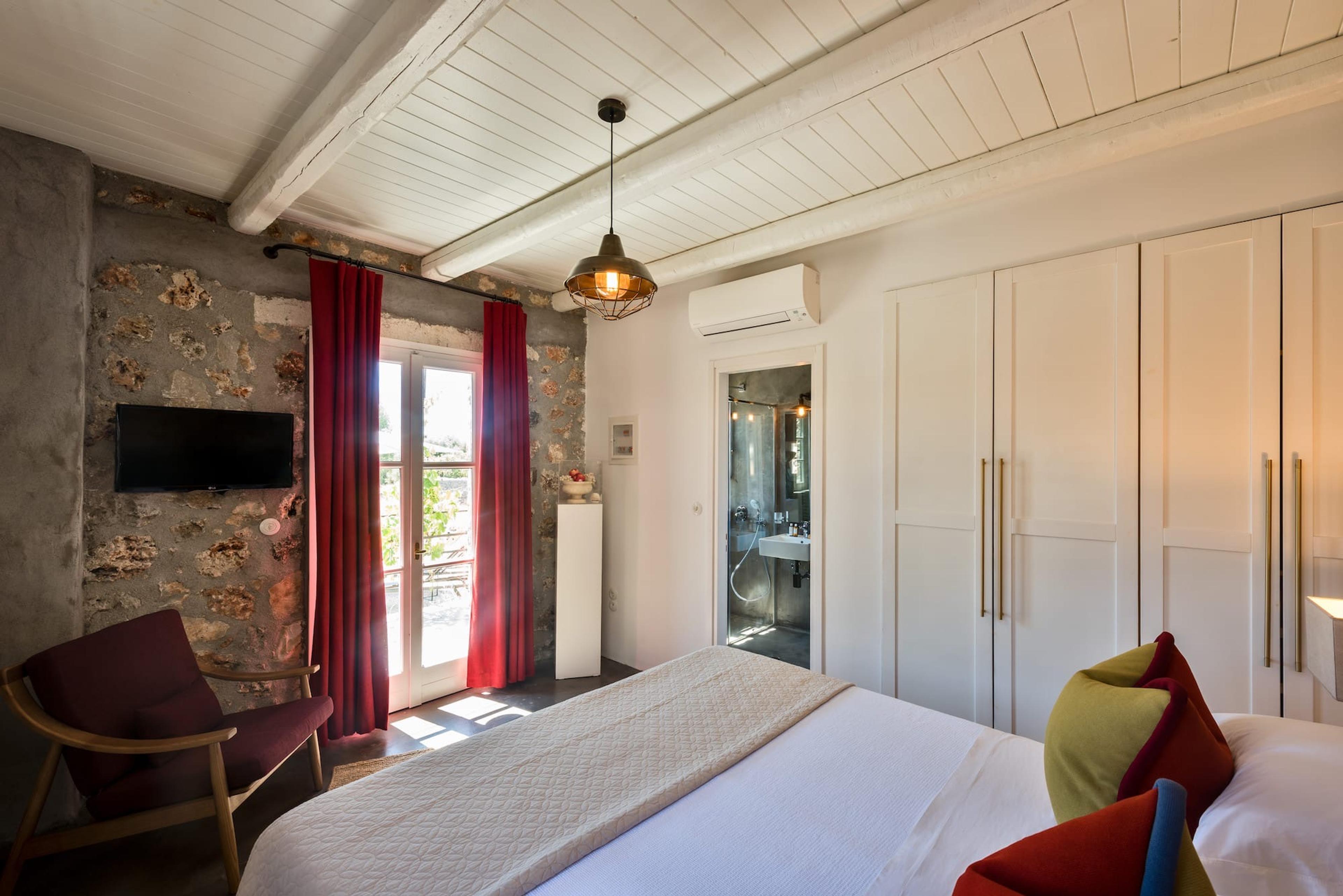 A bedroom with a rough stone wall, a wall-mounted television, a red curtain at the balcony door, and a bed with red, green and blue cushions, with an en-suite bathroom visible through an open door.