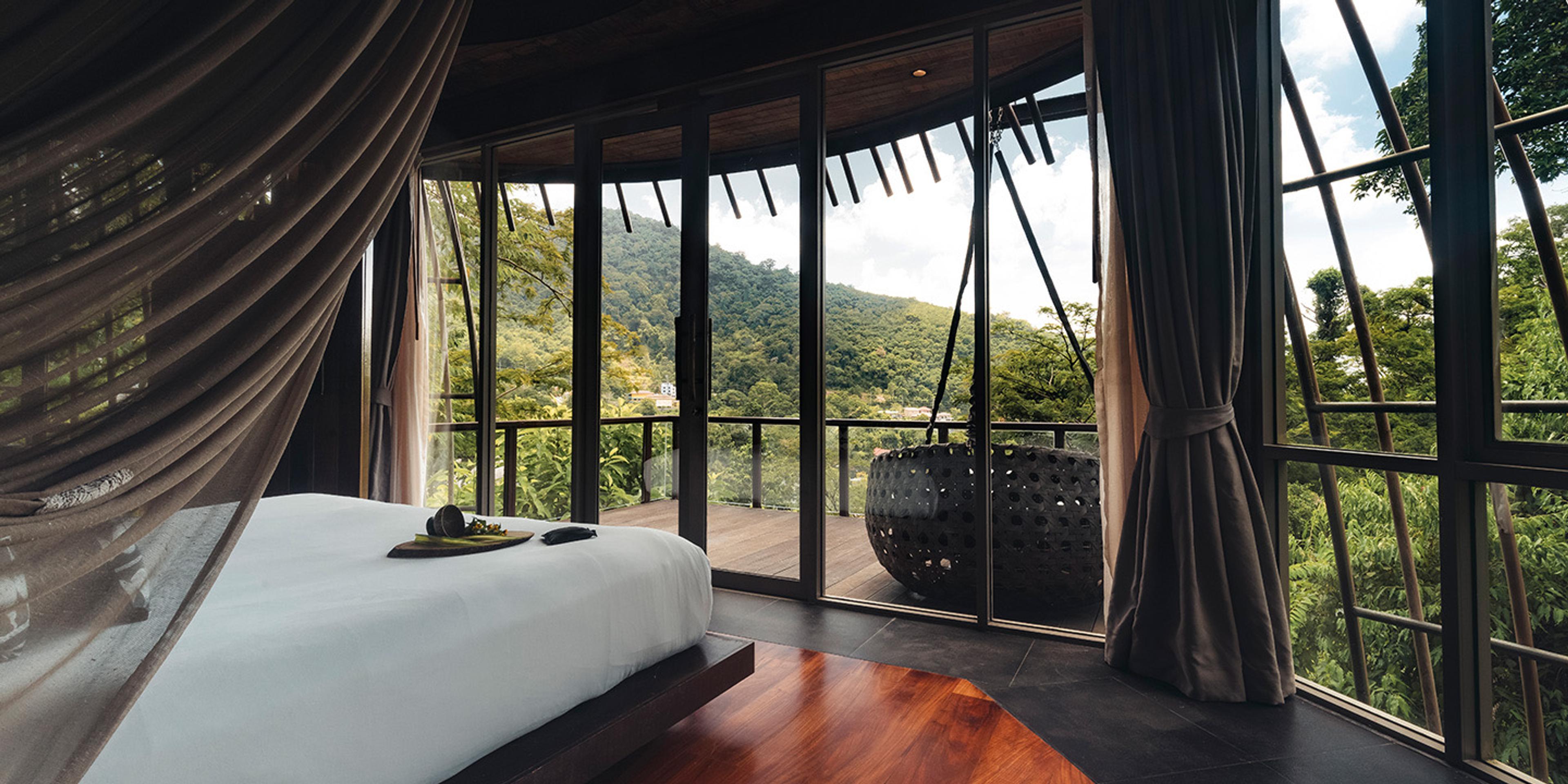 A round, glass-walled bedroom with a bed facing a deck and a wooded mountain view, with a woven hanging chair outside.