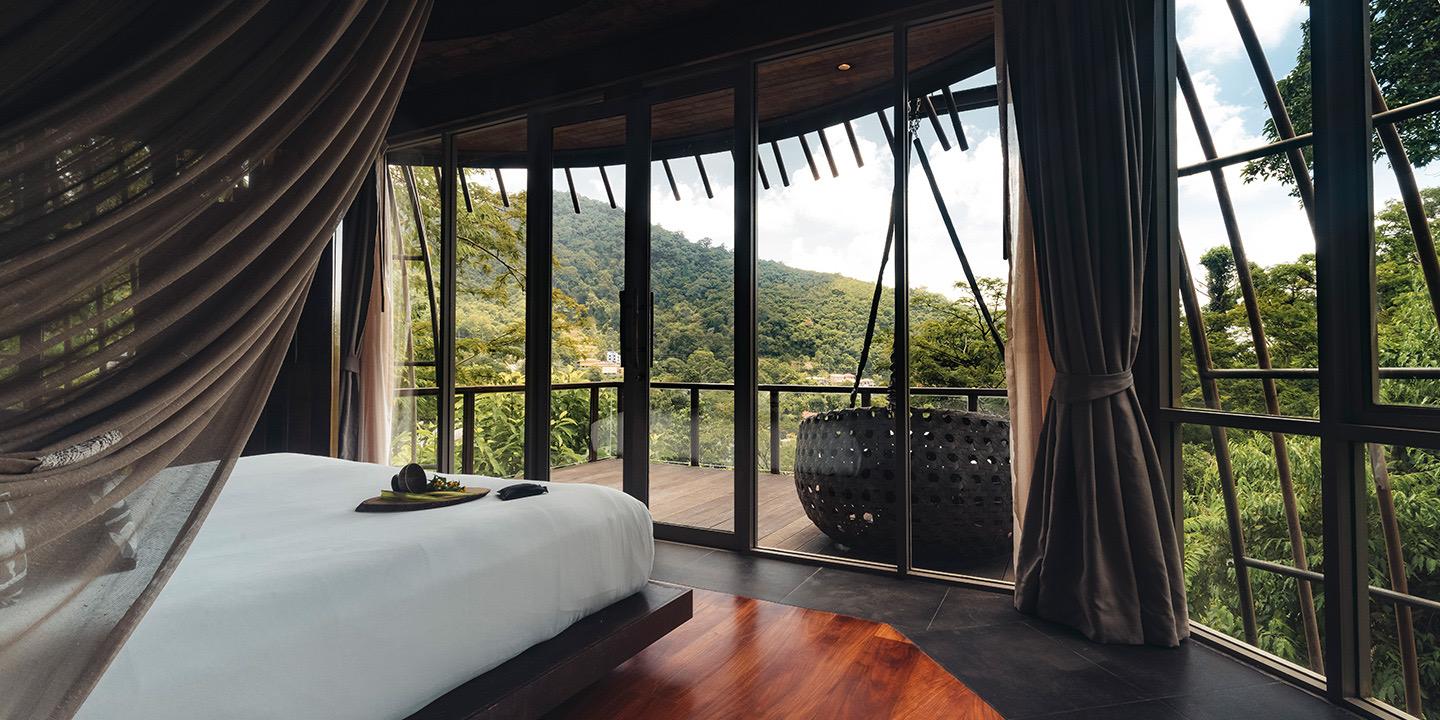 A round, glass-walled bedroom with a bed facing a deck and a wooded mountain view, with a woven hanging chair outside.