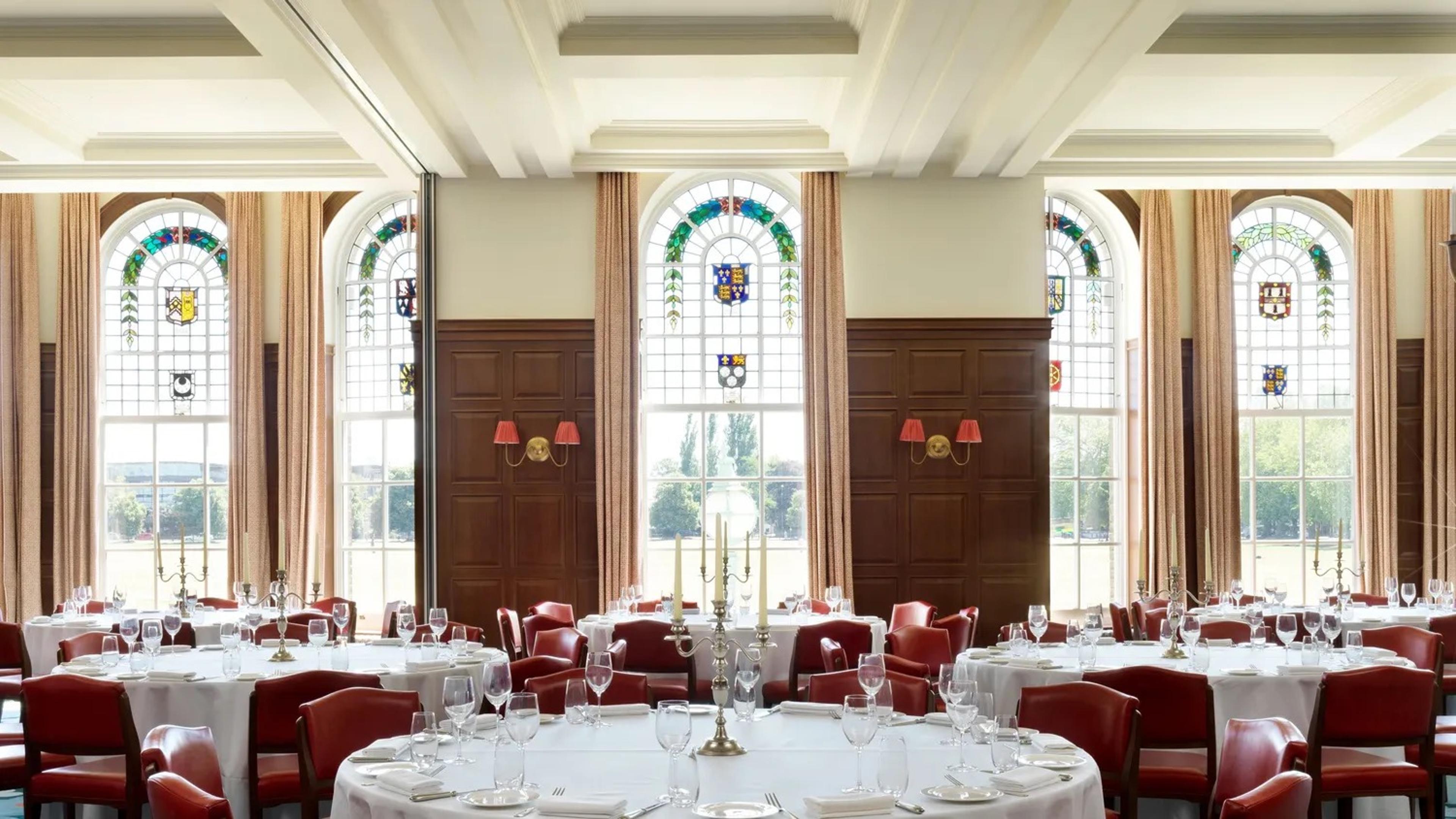 a function room with a high coffered ceiling, tall stained glass windows depicting coats of arms, dark wood panelling, and round tables laid for a formal dinner with red chairs and silver candelabra