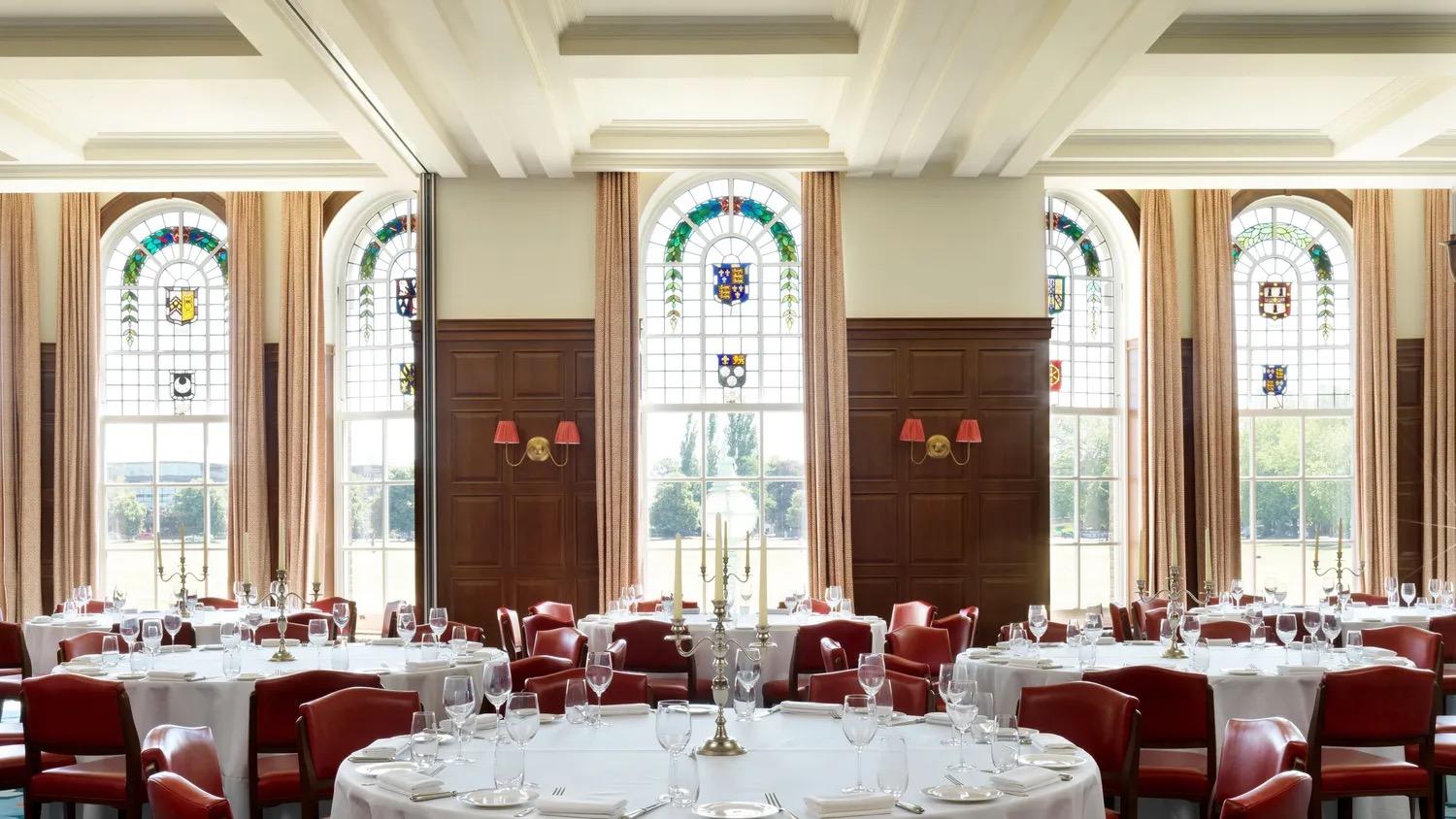 a function room with a high coffered ceiling, tall stained glass windows depicting coats of arms, dark wood panelling, and round tables laid for a formal dinner with red chairs and silver candelabra