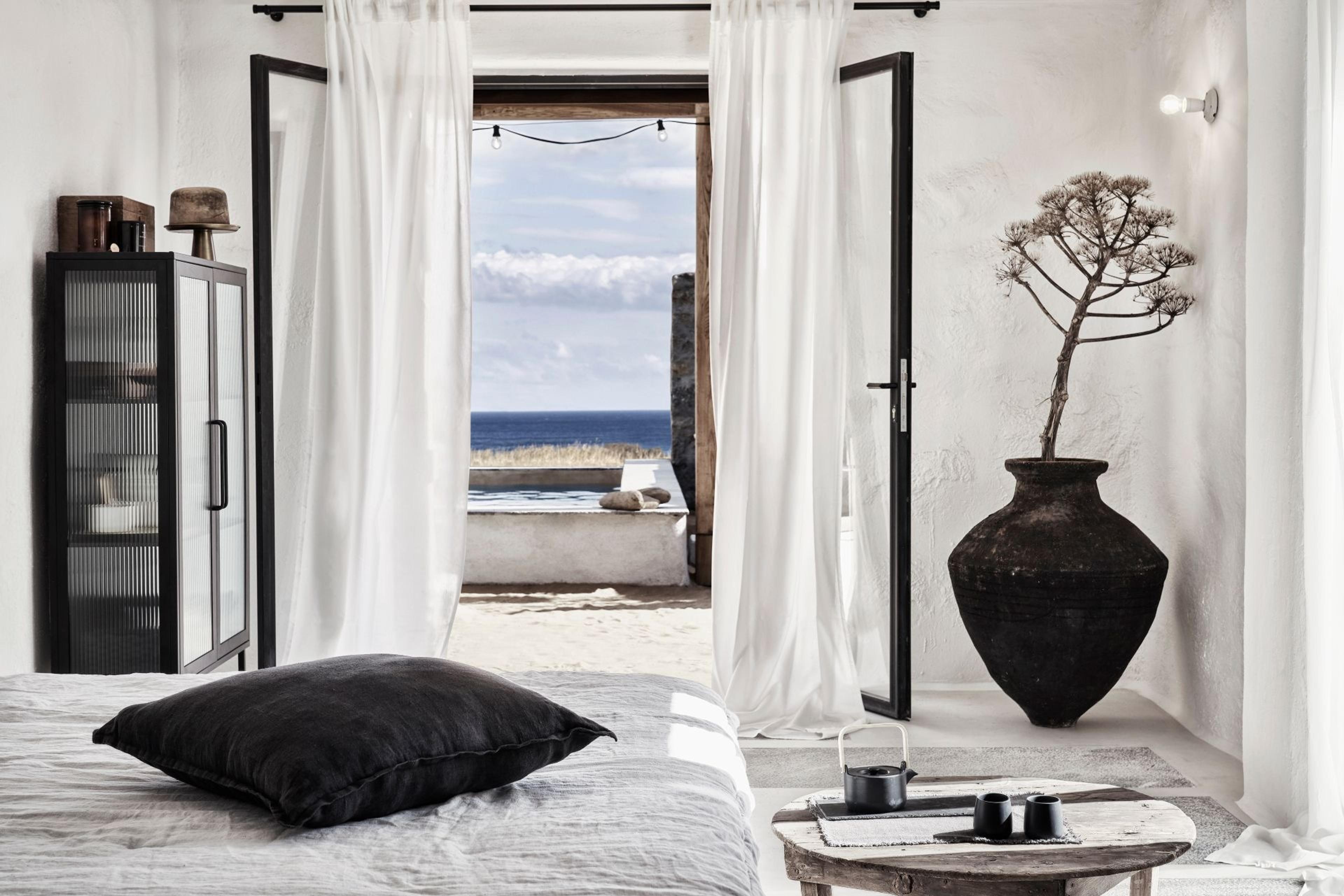 A bedroom with white linen and a black cushion opens through glass doors onto a stone terrace with a plunge pool, the sea visible on the horizon past string lights, and a tall black urn with a dried plant stands beside the doors.