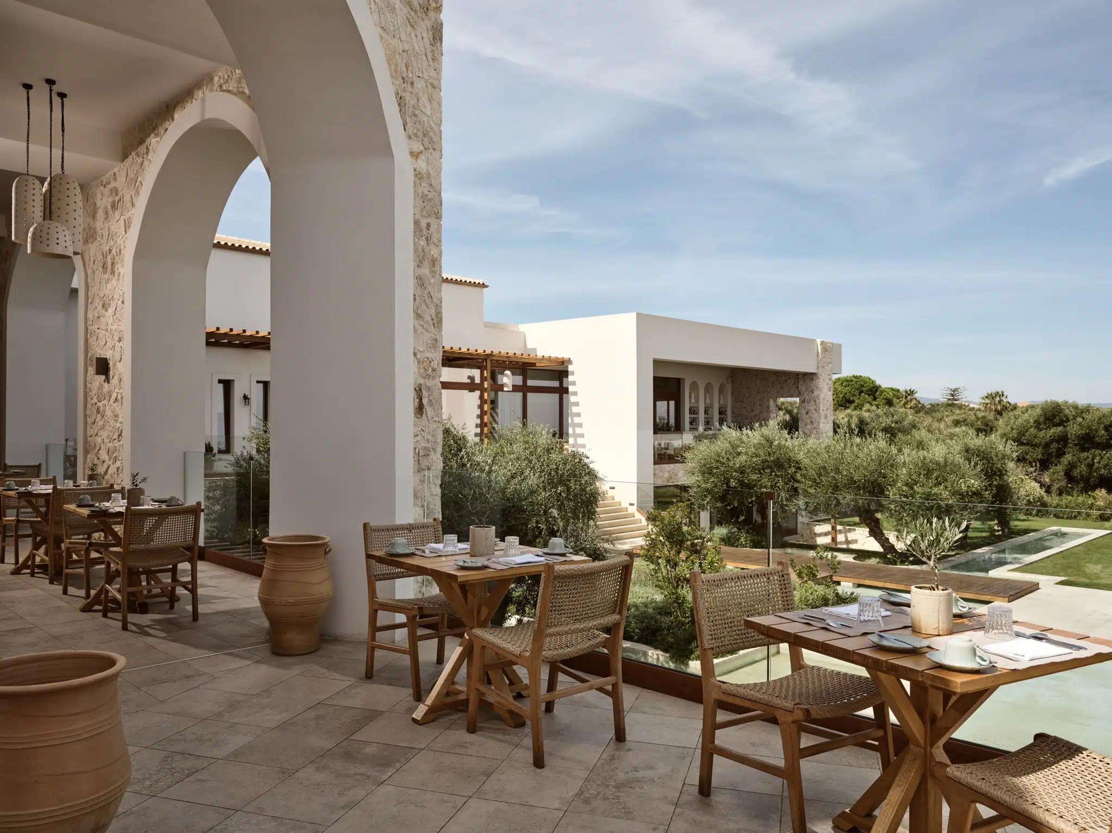 A covered terrace with stone arches and wooden tables set for breakfast, looking over a pool and garden towards the sea.