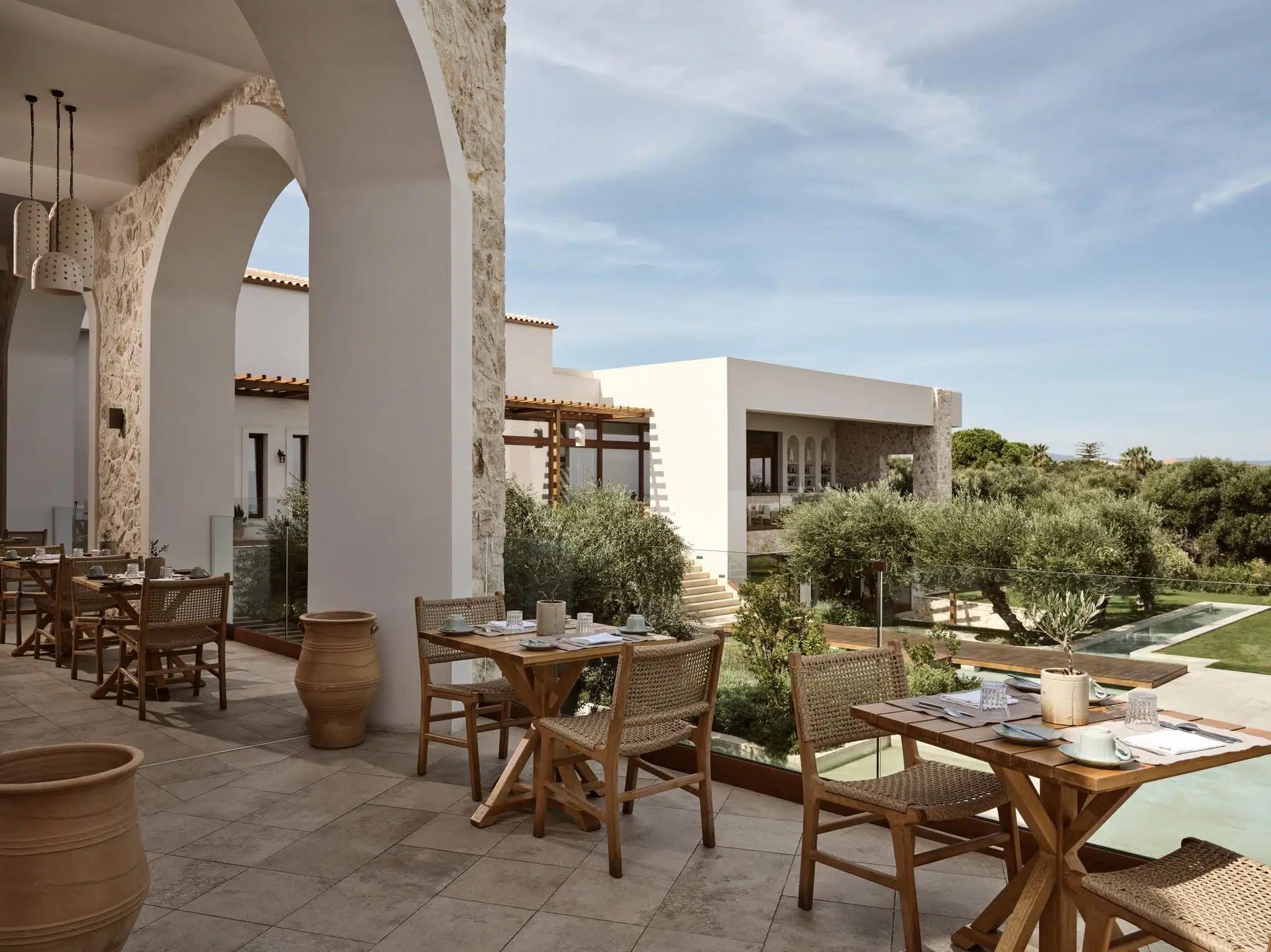 A covered terrace with stone arches and wooden tables set for breakfast, looking over a pool and garden towards the sea.