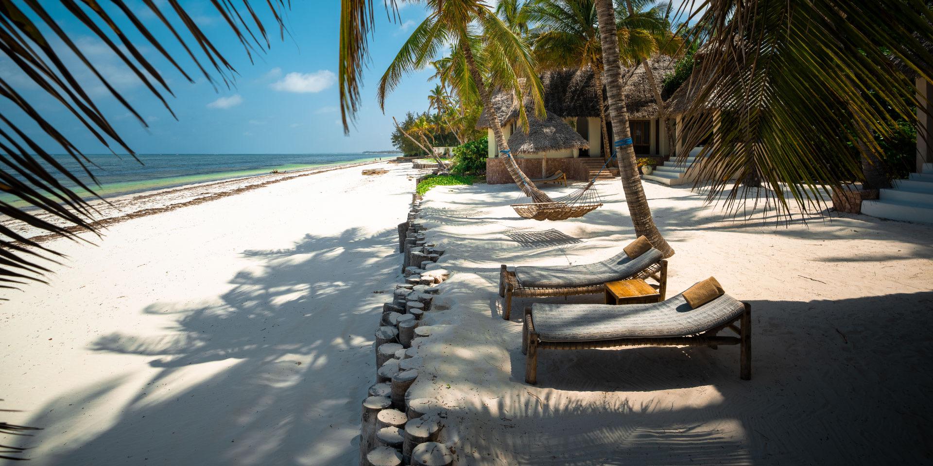 Two shaded loungers and a hammock strung between palms line a white-sand beach in front of thatched villas.