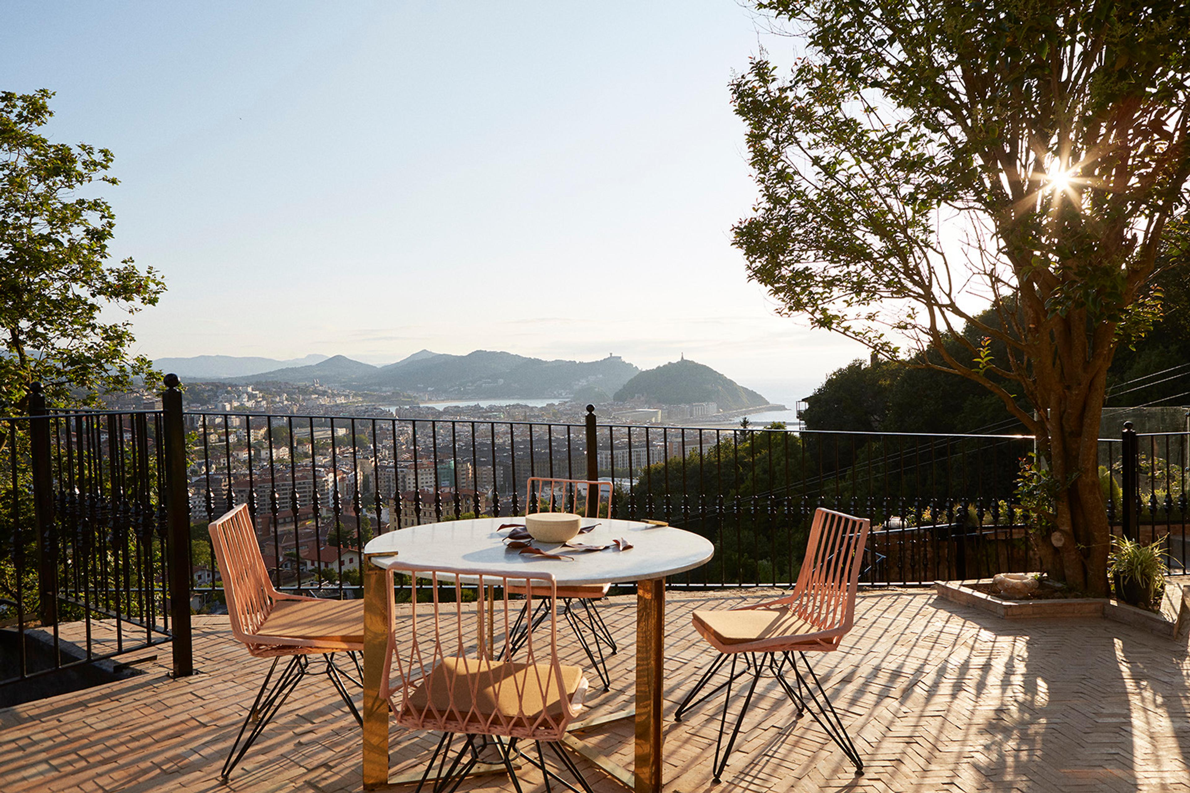 a round table laid with a bowl and set with four chairs on a terrace under a tree, overlooking the bay, the town and the hills with the sun breaking through the branches
