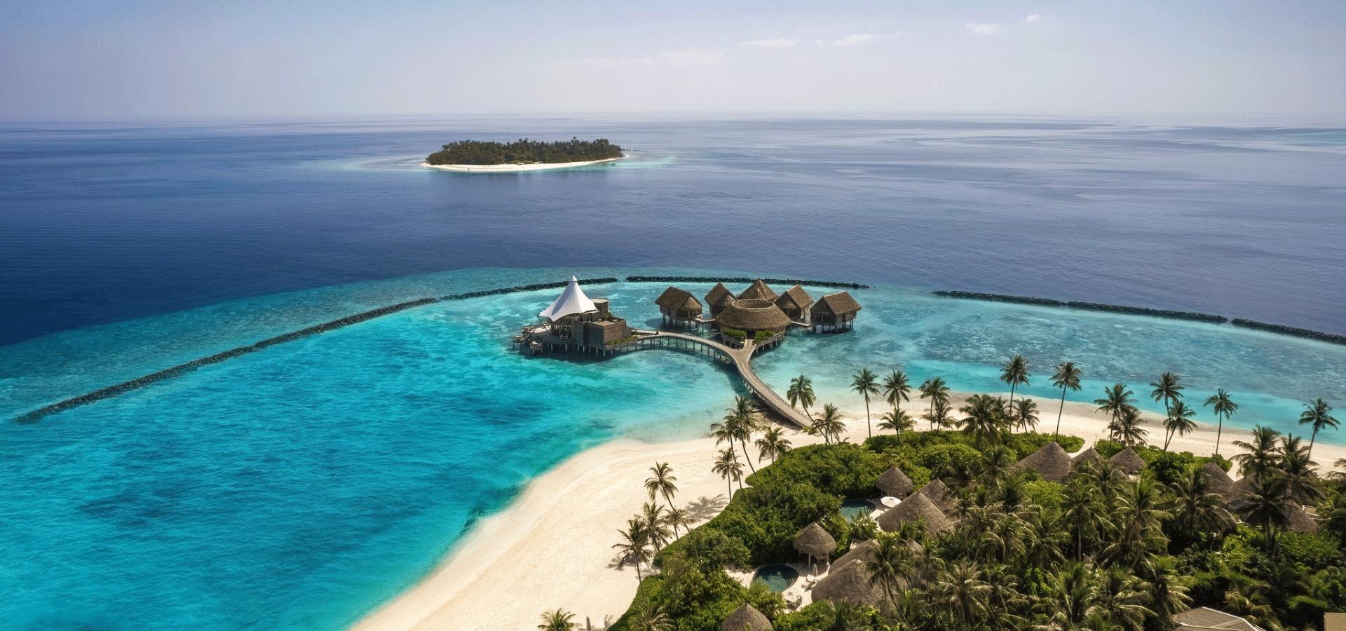 A wide aerial view of the island resort surrounded by reef and turquoise water, with a smaller uninhabited island visible on the horizon.