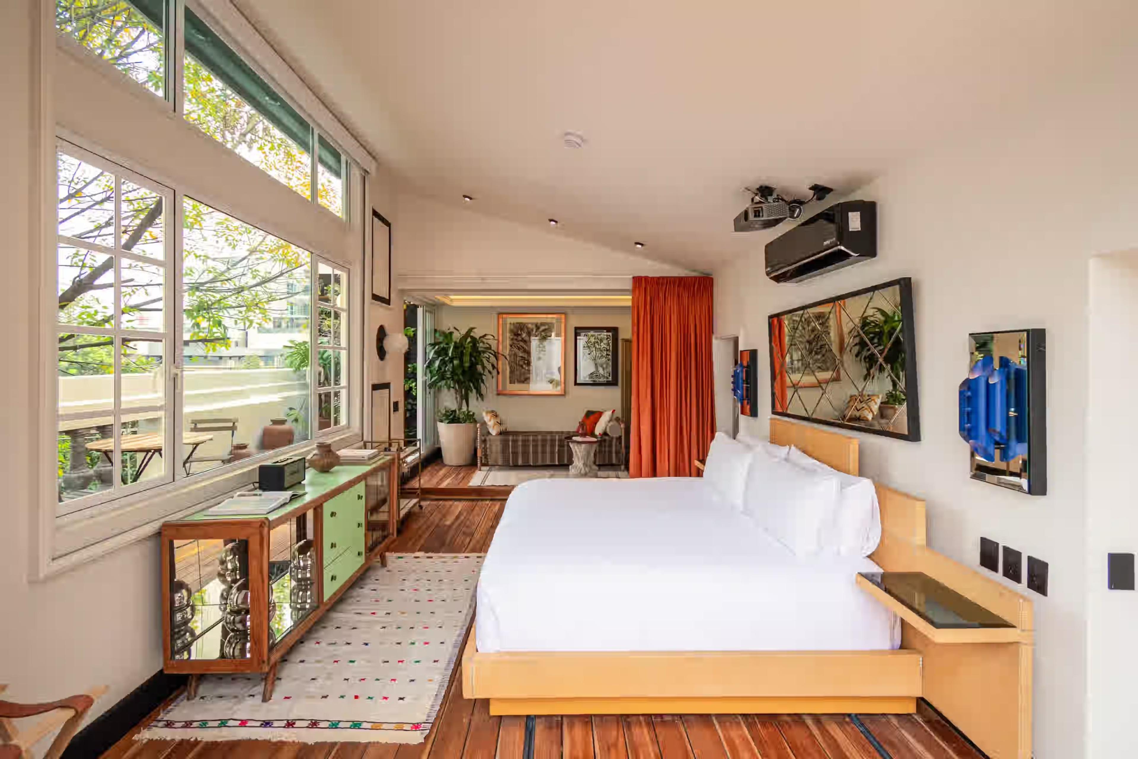 A bedroom has a floor-to-ceiling window onto leafy trees, a green dresser, and a sitting area with an orange curtain beyond.