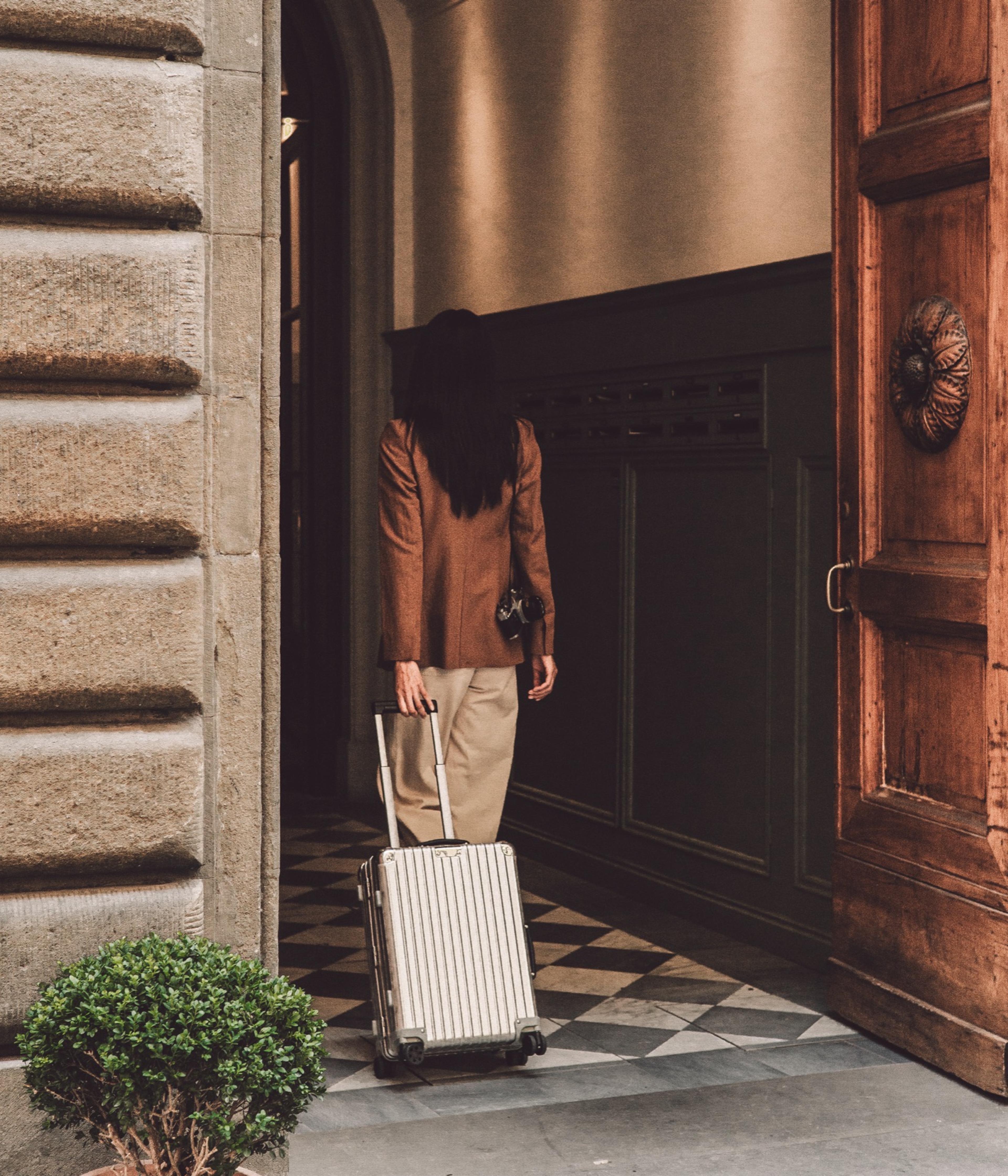 A woman seen from behind wheels a silver suitcase across a black and white checkerboard floor into a wood-paneled entrance hall, past a carved wooden door and a potted plant.